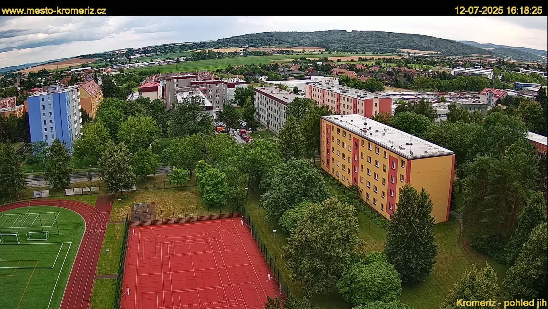 An aerial view shows apartment buildings, green spaces with trees, and a red athletic field, all under a sunny sky.