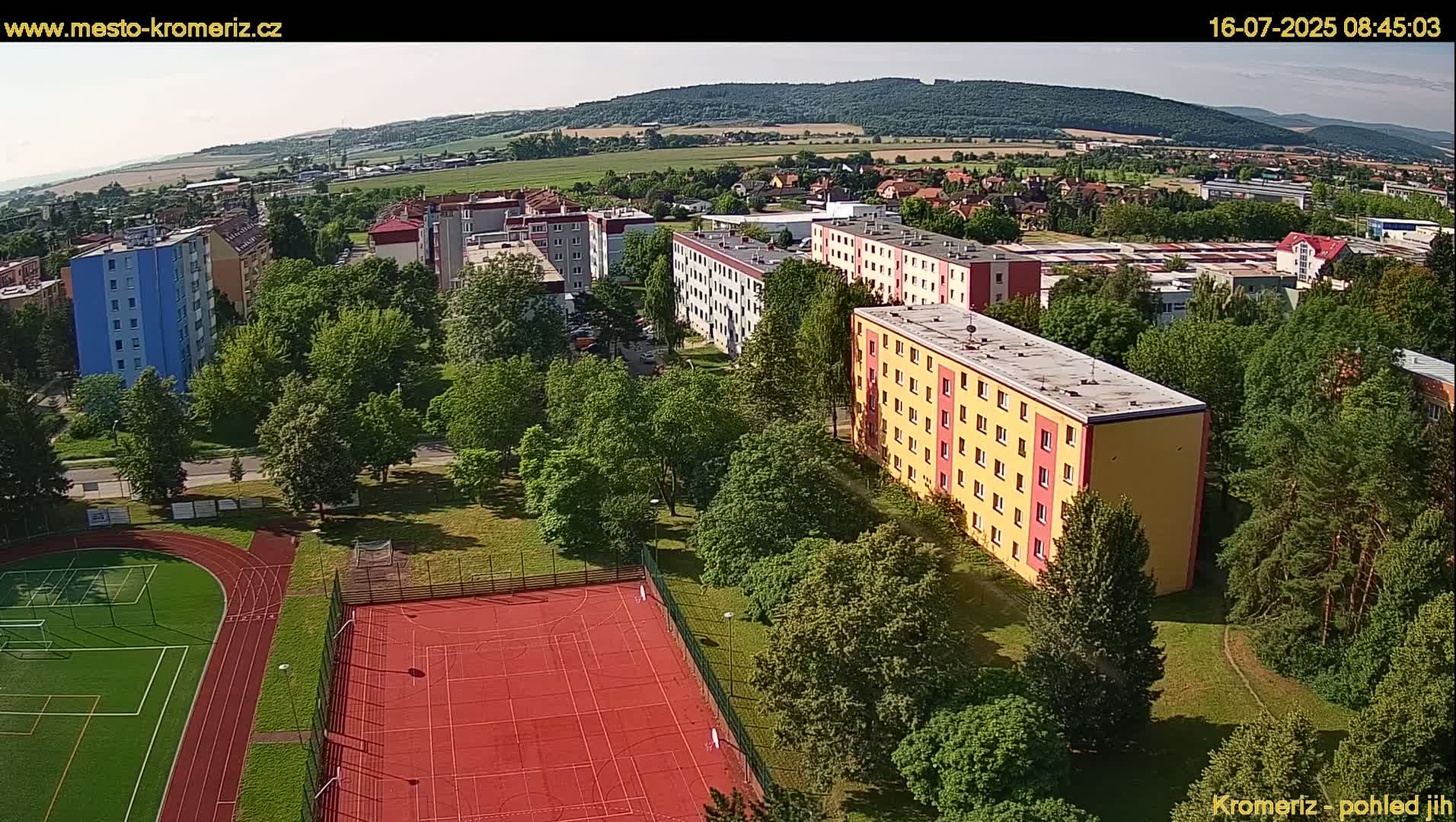 An aerial view shows a residential area with multi-story apartment buildings, green spaces including a park and sports field, and rolling hills in the background under a clear sky.