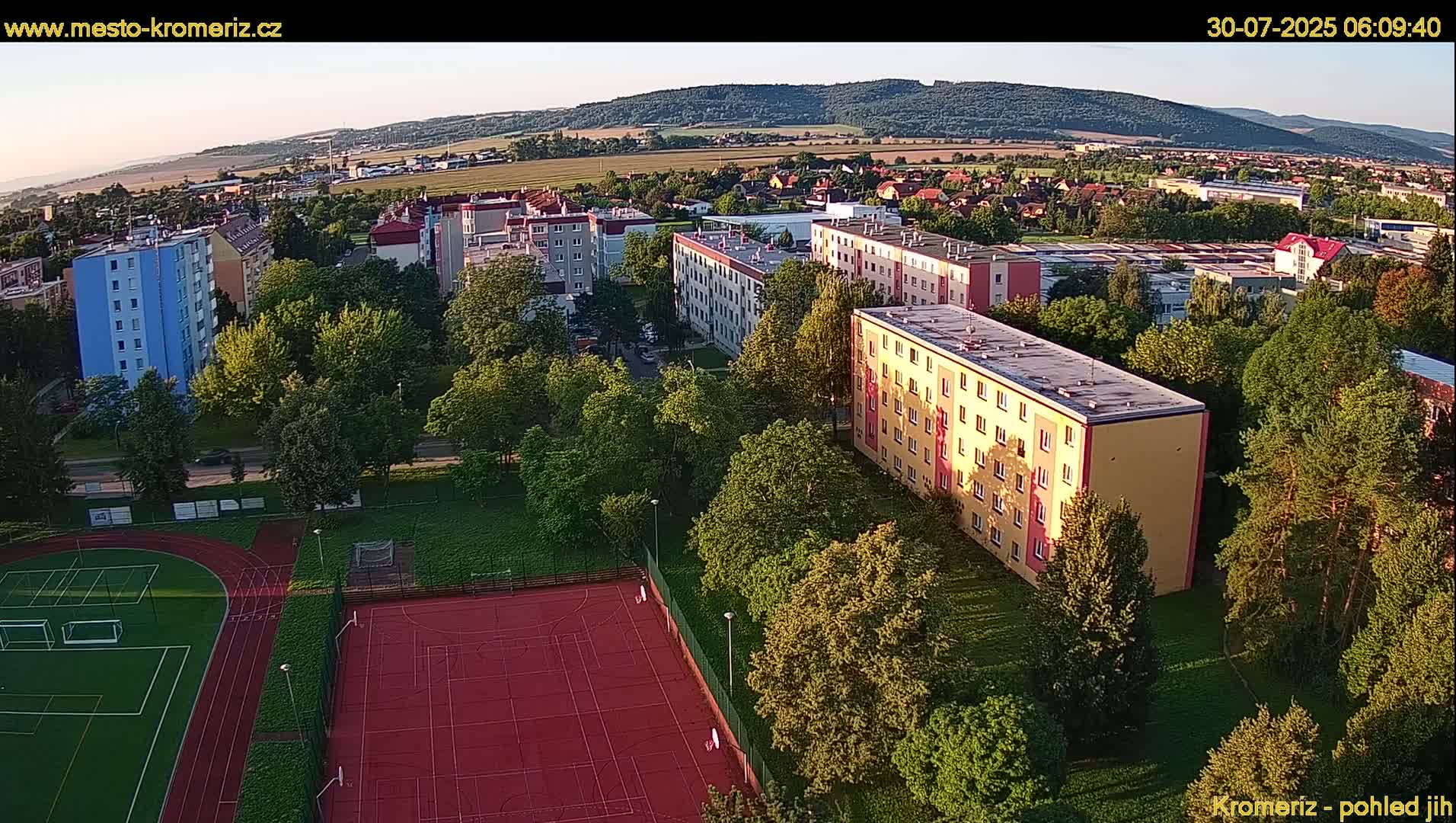An aerial view shows apartment buildings surrounded by lush green trees and a red sports court, under a mostly sunny sky.