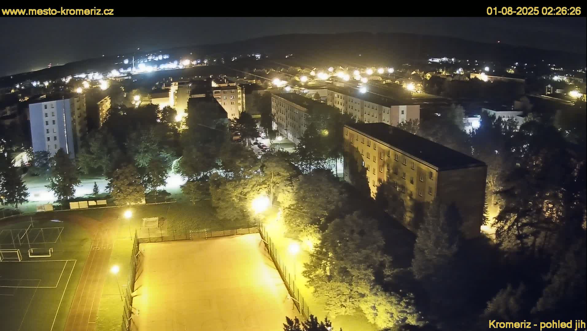 A nighttime aerial view of a residential area with illuminated buildings, trees, and a sports field.