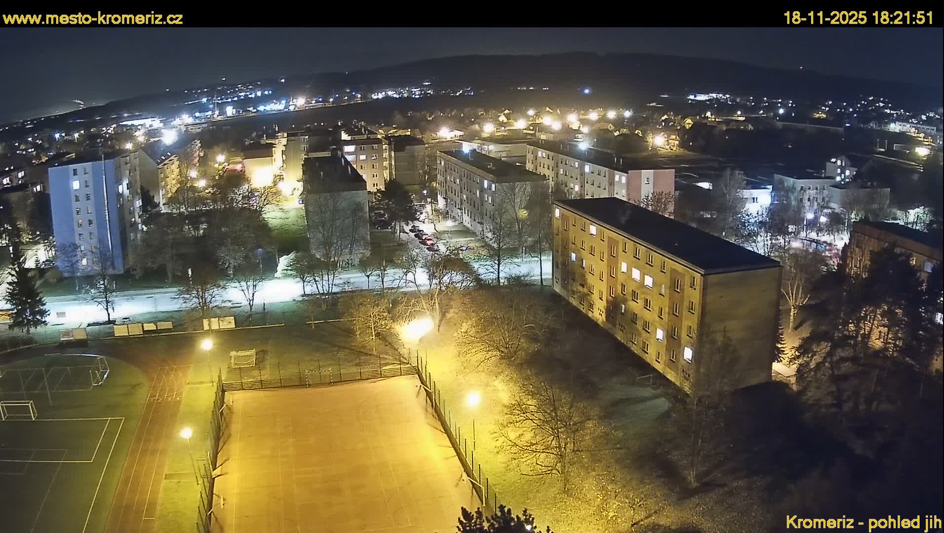 An aerial view shows a residential area with multi-story buildings surrounded by green trees and athletic fields under a mostly sunny sky.