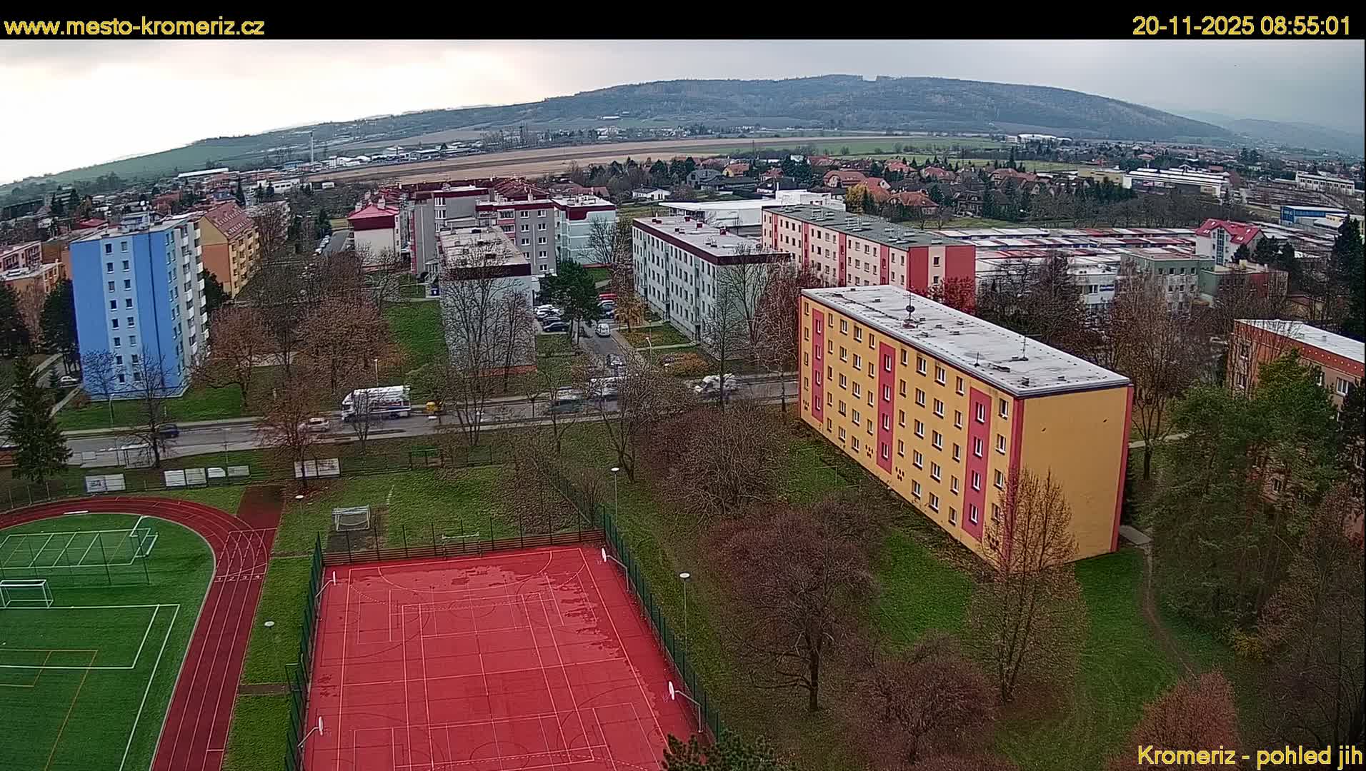 An aerial view captures a residential area with colorful apartment buildings, dormant trees, sports fields, and roads under a cloudy, overcast sky, with rolling hills visible in the distance.