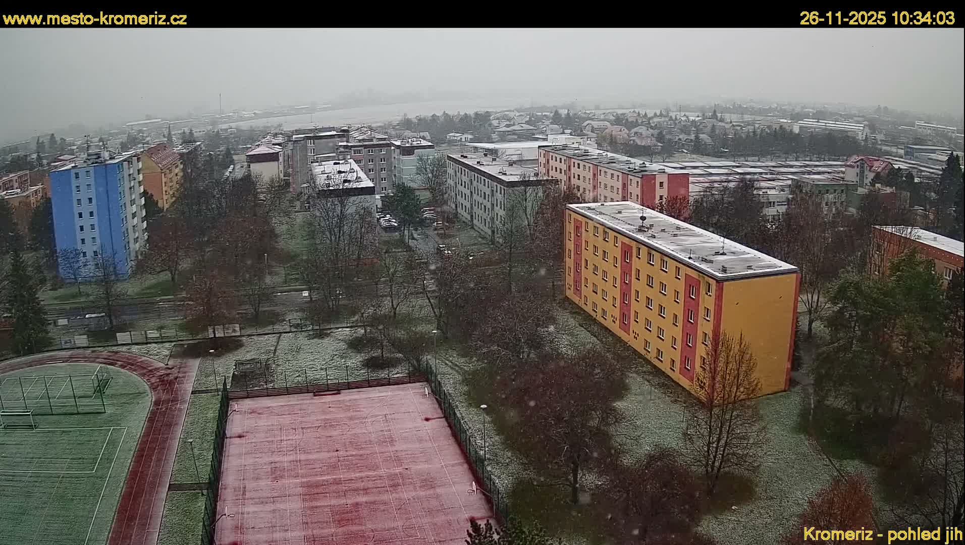 I see a wintry residential landscape featuring multiple apartment buildings, sports fields, and bare trees lightly covered in snow, with active snowfall under a gray, overcast sky.