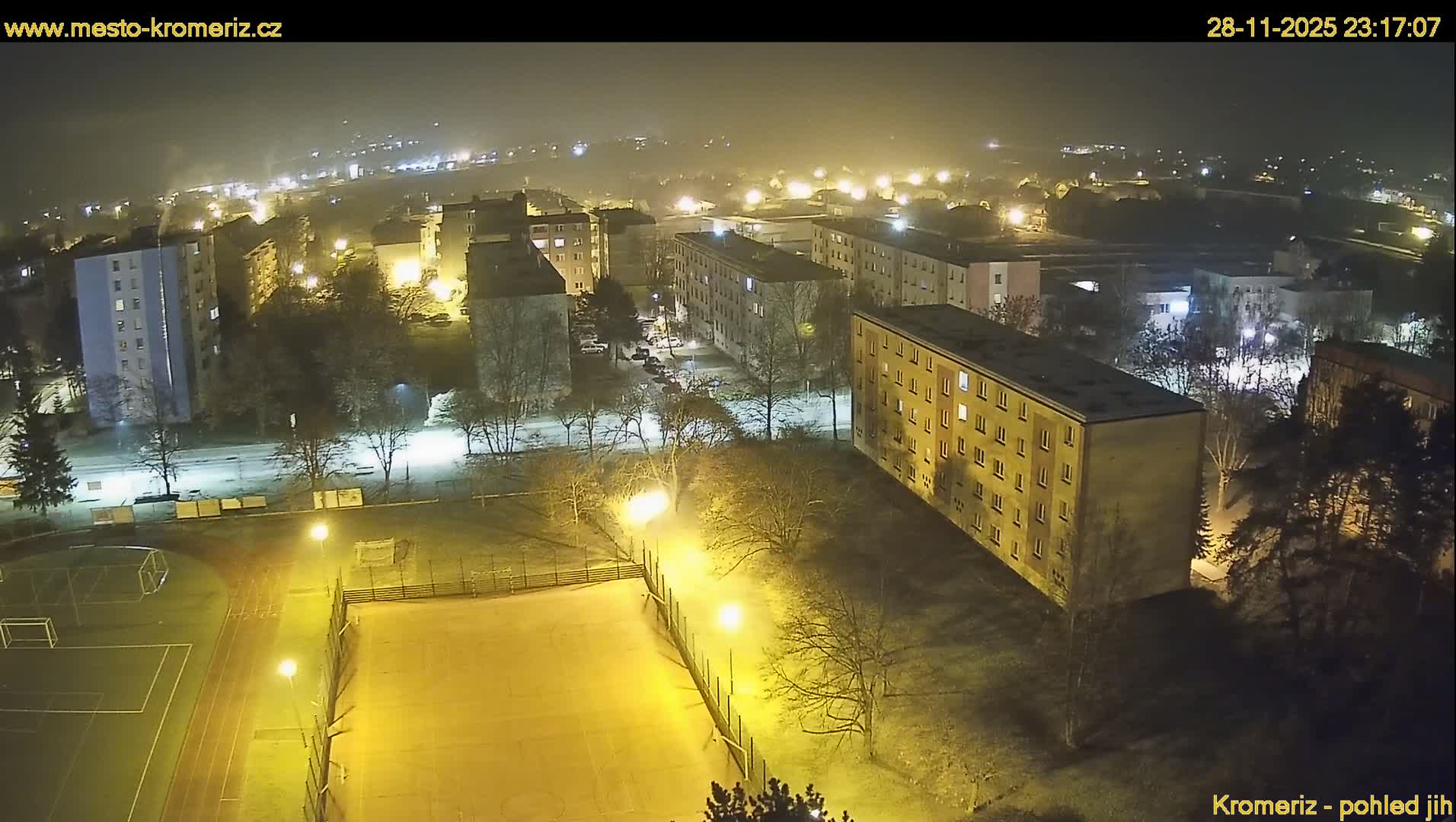 From an elevated perspective, a hazy winter night illuminates a residential area featuring numerous apartment buildings, bare trees, and a sports complex, with light ground cover suggesting frost or a thin layer of snow.