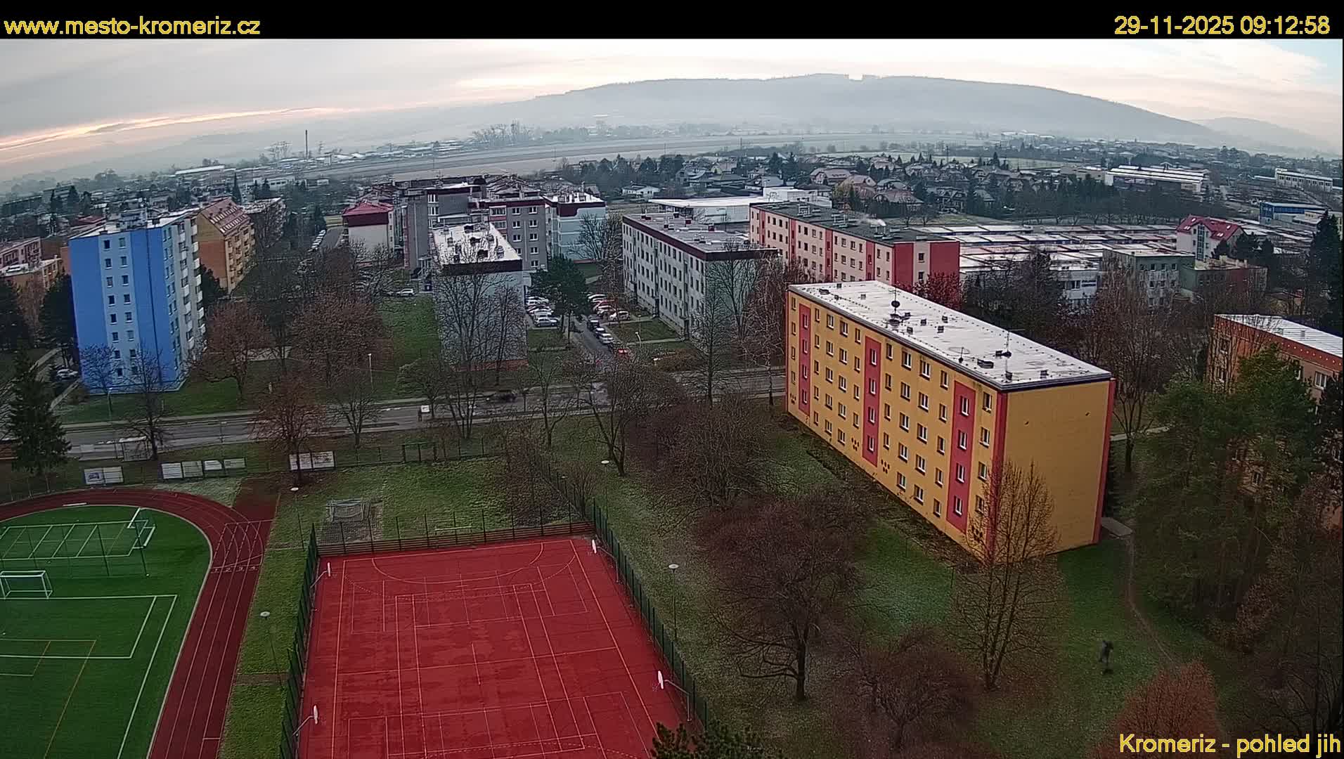 An elevated view captures a residential area on a hazy, overcast day, featuring multi-story apartment blocks in various colors, a red running track and an adjacent sports court, bare trees, and a distant cityscape fading into misty hills.