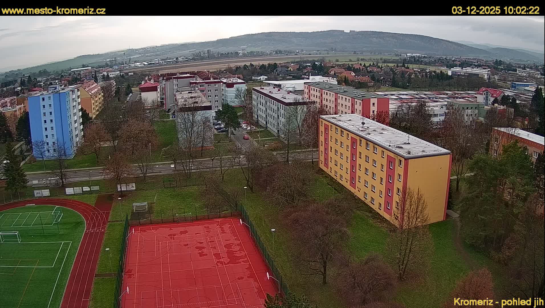 An aerial view reveals a sprawling residential area featuring colorful apartment blocks, sports fields, and bare trees under an overcast sky, with distant hills visible on a damp, likely late autumn or winter day.