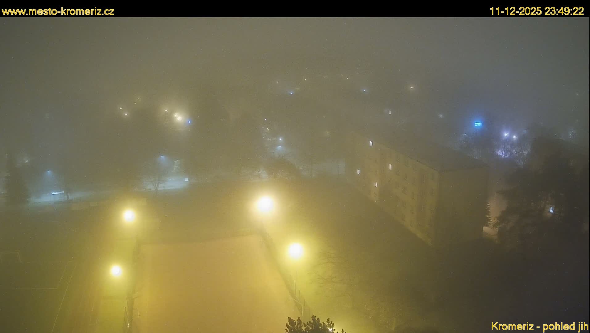 A thick fog obscures a nighttime urban scene, illuminated by glowing streetlights and scattered building lights, with a sports court faintly visible in the foreground.