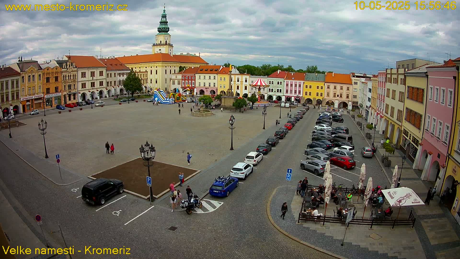 A large town square with colorful buildings, parked cars, and a few people, under a partly cloudy sky.