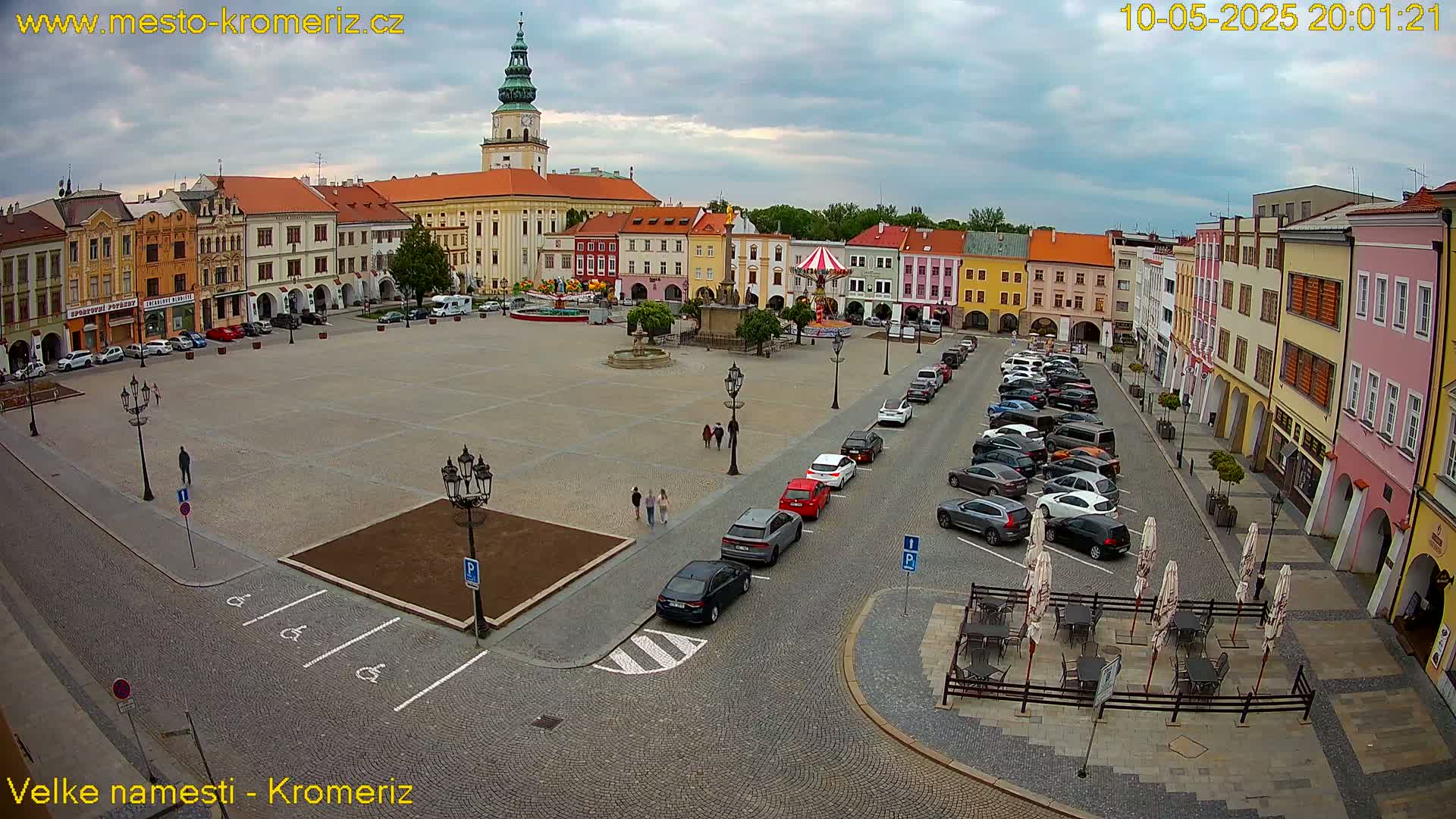 A large town square with colorful buildings surrounding it, cars parked along the sides, and a few people walking, under an overcast sky.
