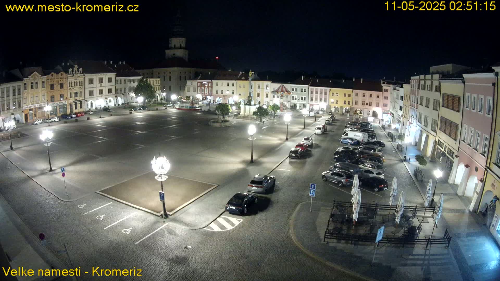 A nighttime view of a town square with parked cars, streetlights, and buildings surrounding a central open space.