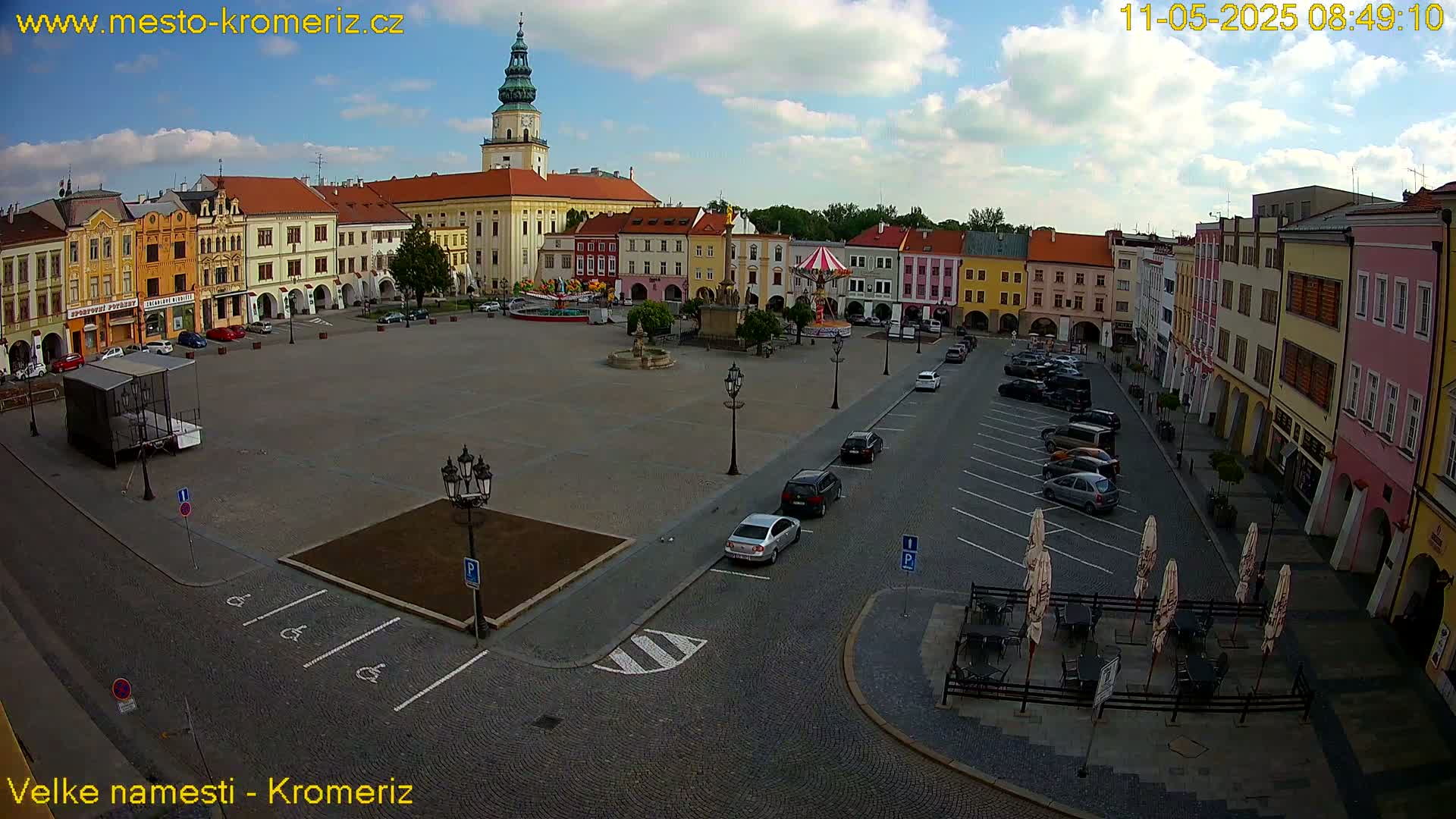 A town square with colorful buildings, a carousel, and parked cars under a partly cloudy sky.