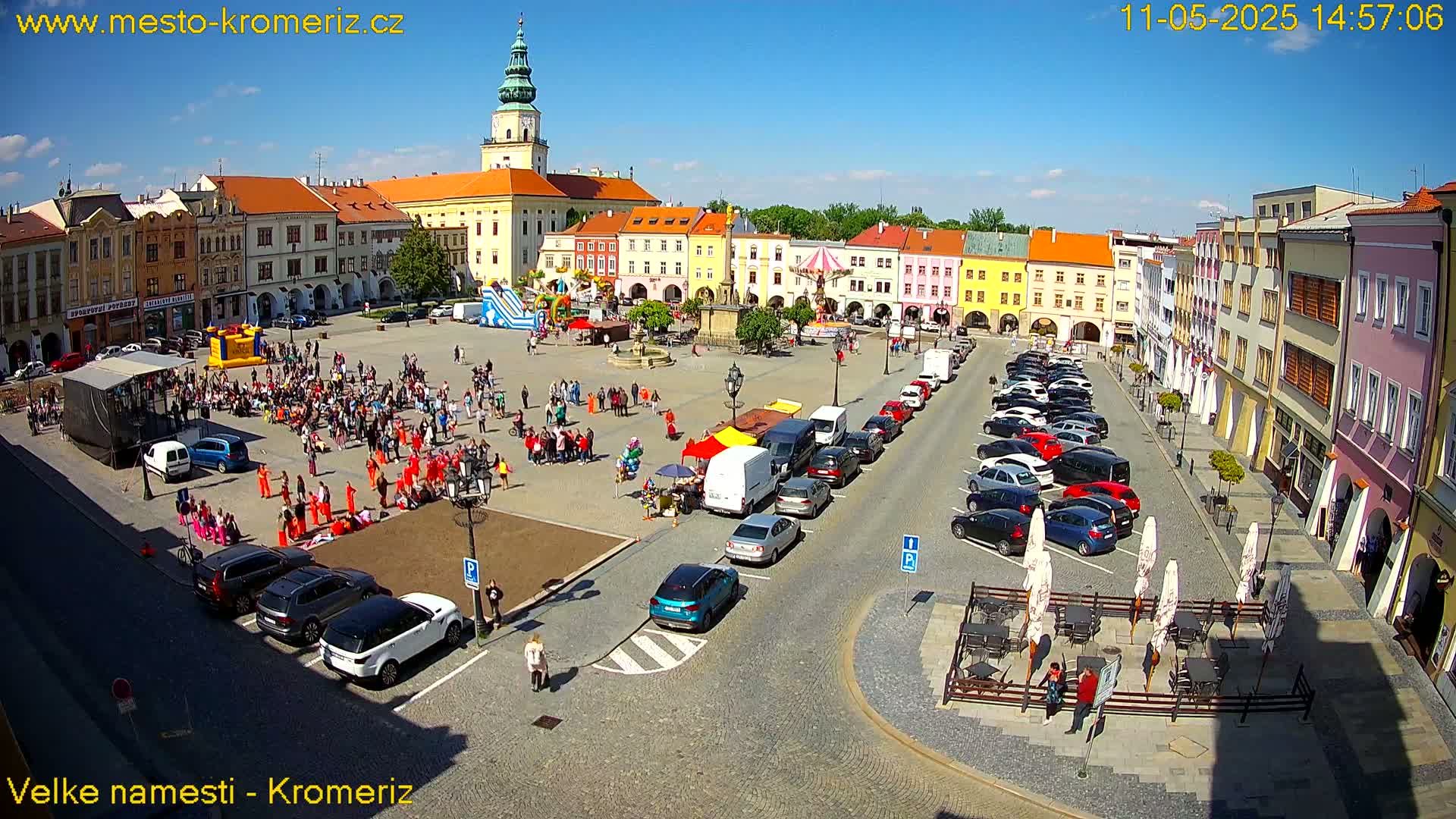 A sunny town square with colorful buildings, a crowd of people, parked cars, and outdoor seating areas.