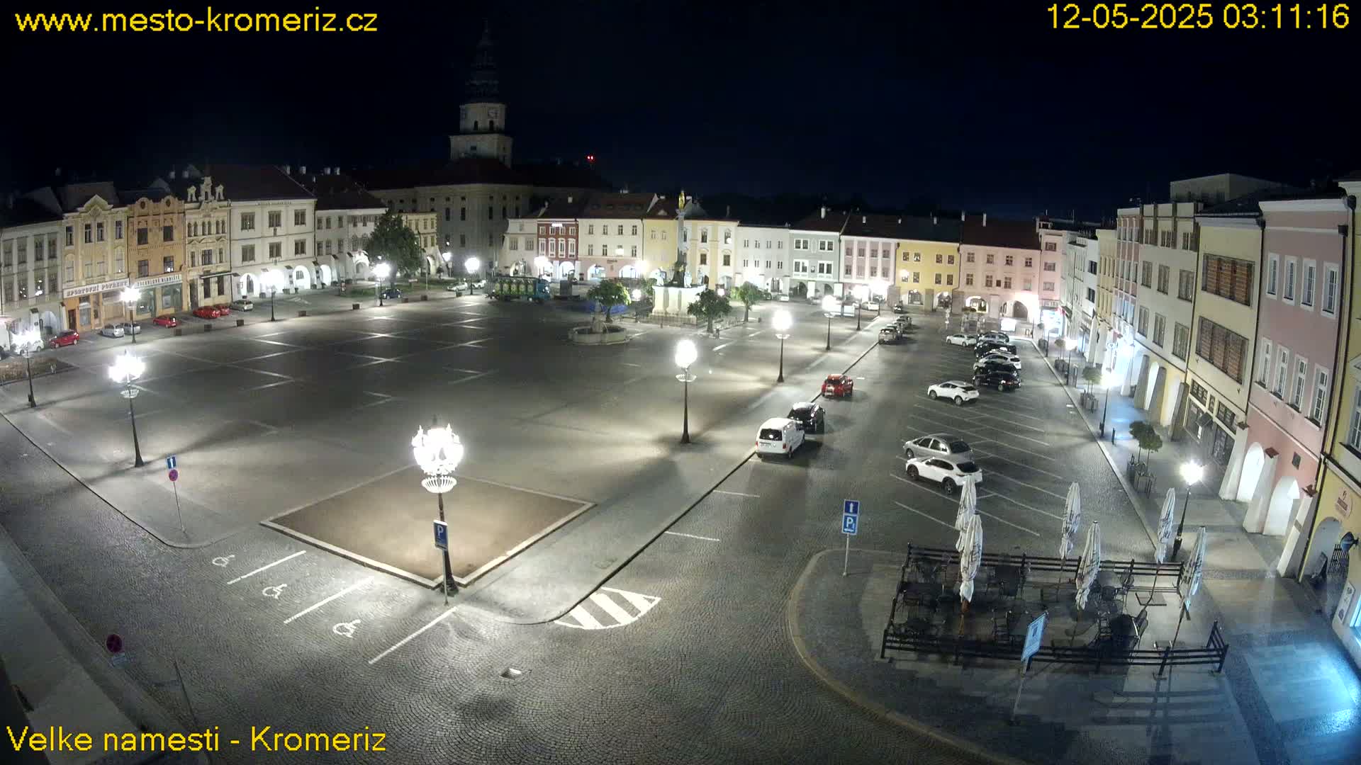A town square at night, illuminated by streetlights, with several cars parked and outdoor seating at a cafe.