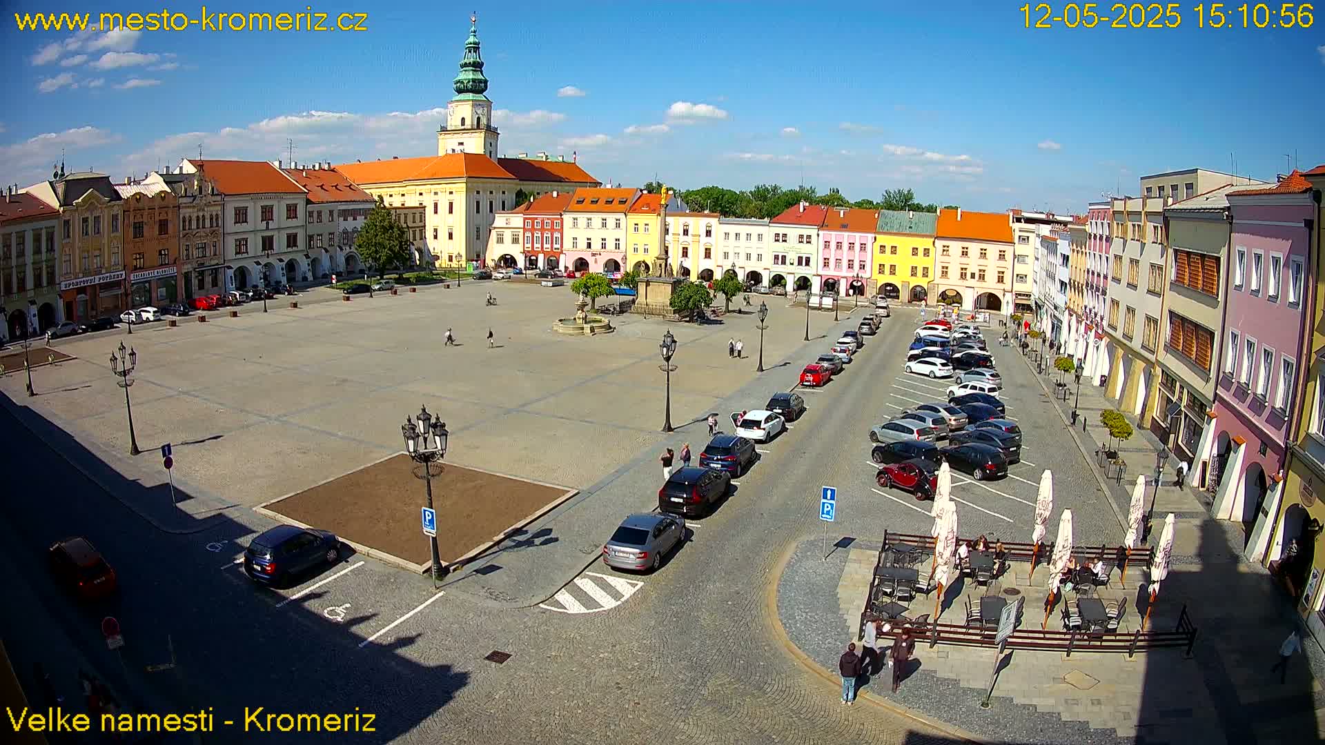 A large town square with colorful buildings, a fountain, parked cars, and outdoor seating under umbrellas on a sunny day.