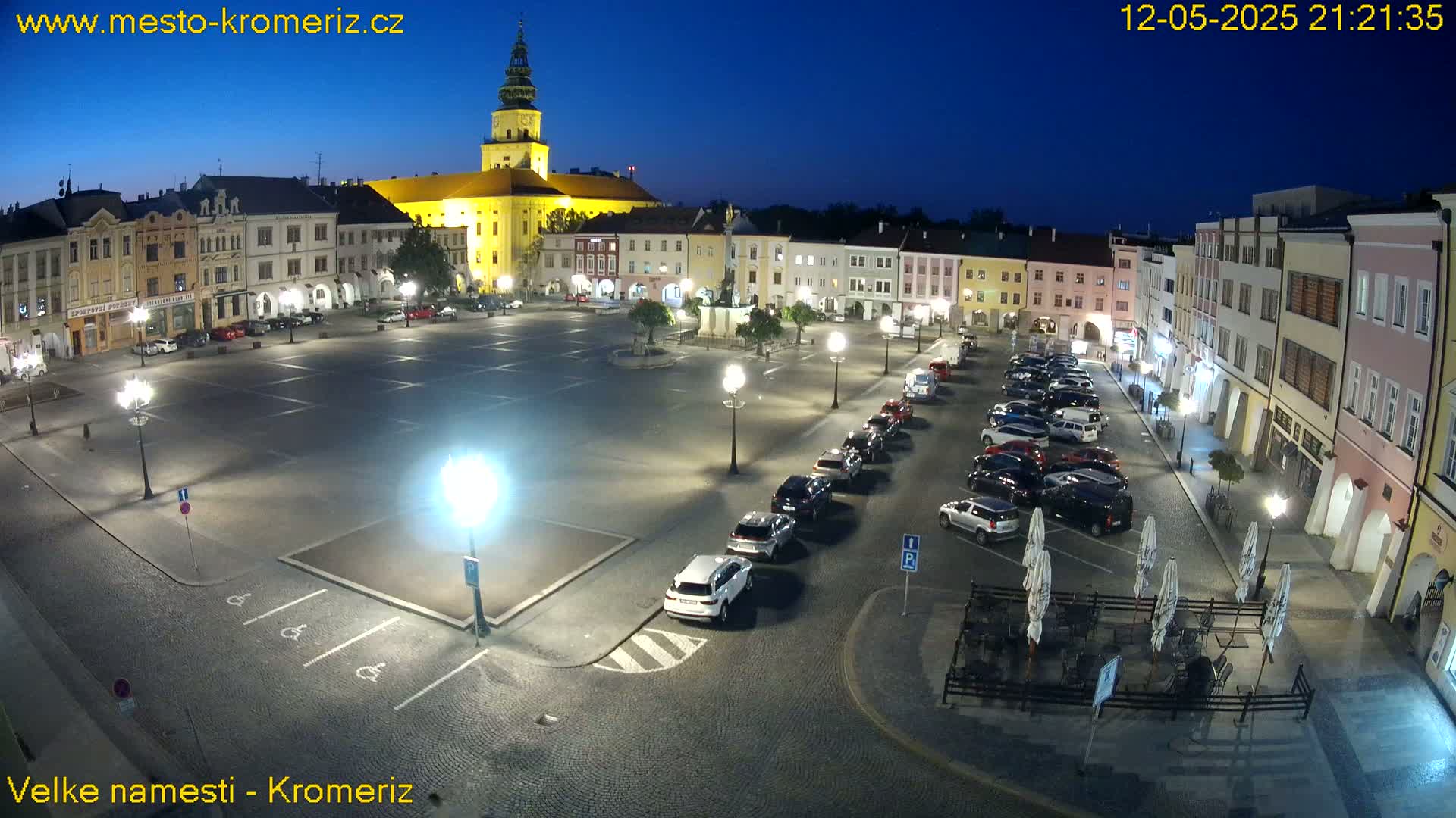 A large, empty town square at night, illuminated by streetlights, is surrounded by historic buildings and has cars parked along the edges.