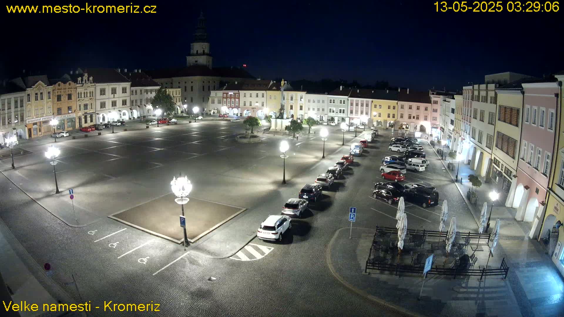 A nighttime view of a town square with cars parked along the sides and a few outdoor tables and chairs, under a clear, dark sky.