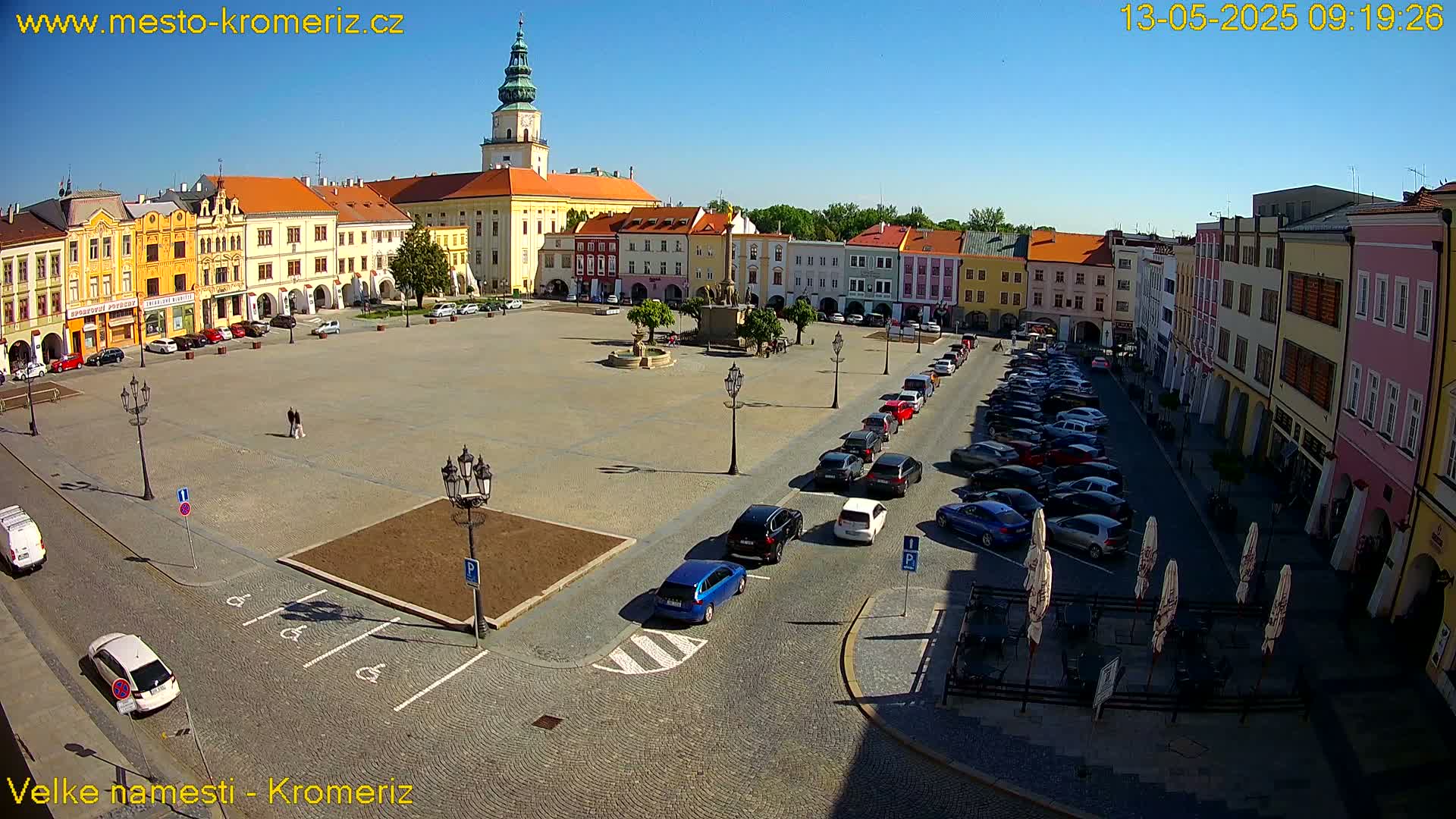 A large town square with colorful buildings surrounding it, cars parked along the edges, and a fountain in the center, under sunny skies.