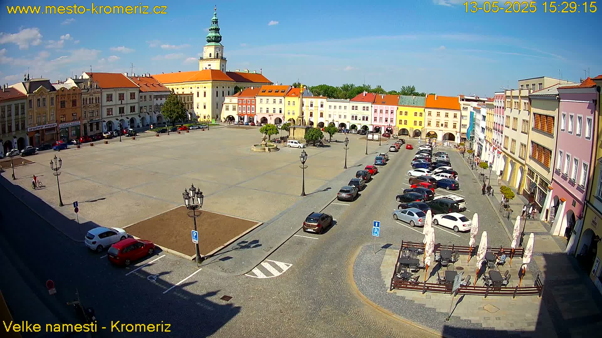 A large town square with colorful buildings, a fountain, and parked cars under a sunny sky.
