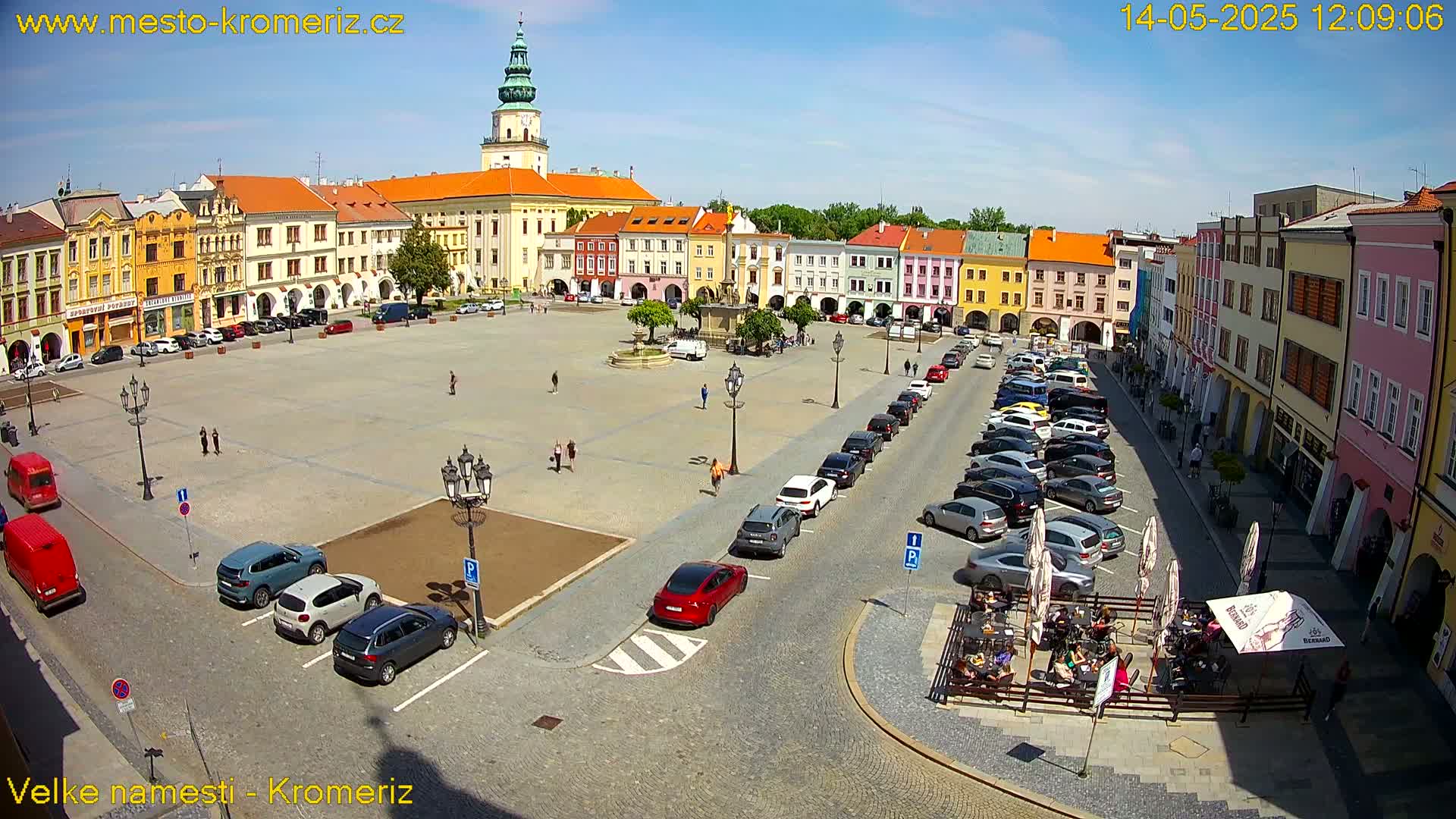 A large town square with colorful buildings surrounding it, numerous cars parked along the sides, and a few pedestrians, all under a clear sunny sky.