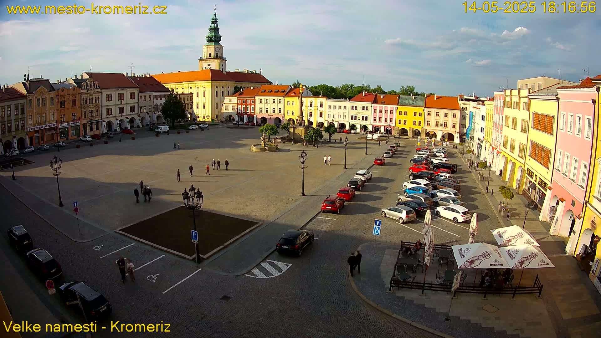 A large town square with colorful buildings surrounding it, under a partly cloudy sky, is populated with several parked cars and a few pedestrians.