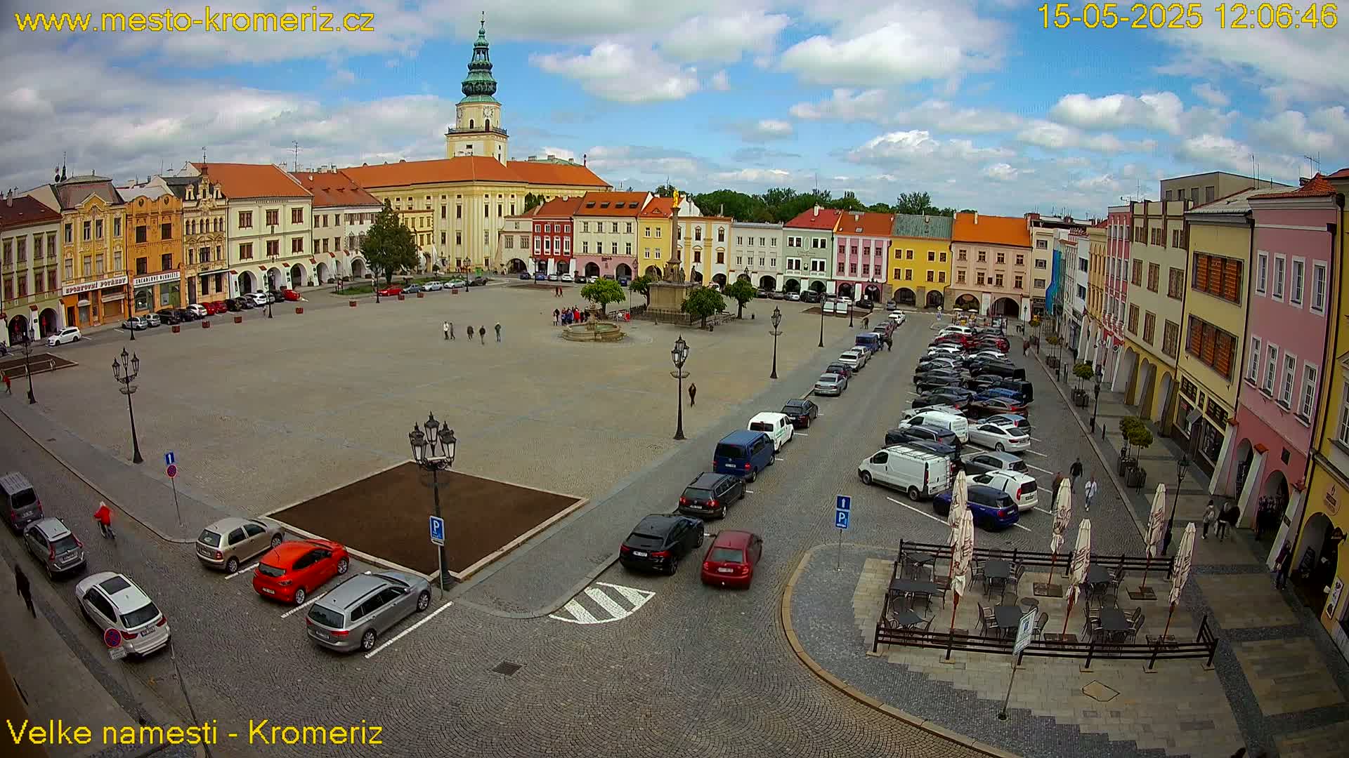A town square with colorful buildings surrounding a paved area, cars parked along the sides, and a few people walking around under a partly cloudy sky.