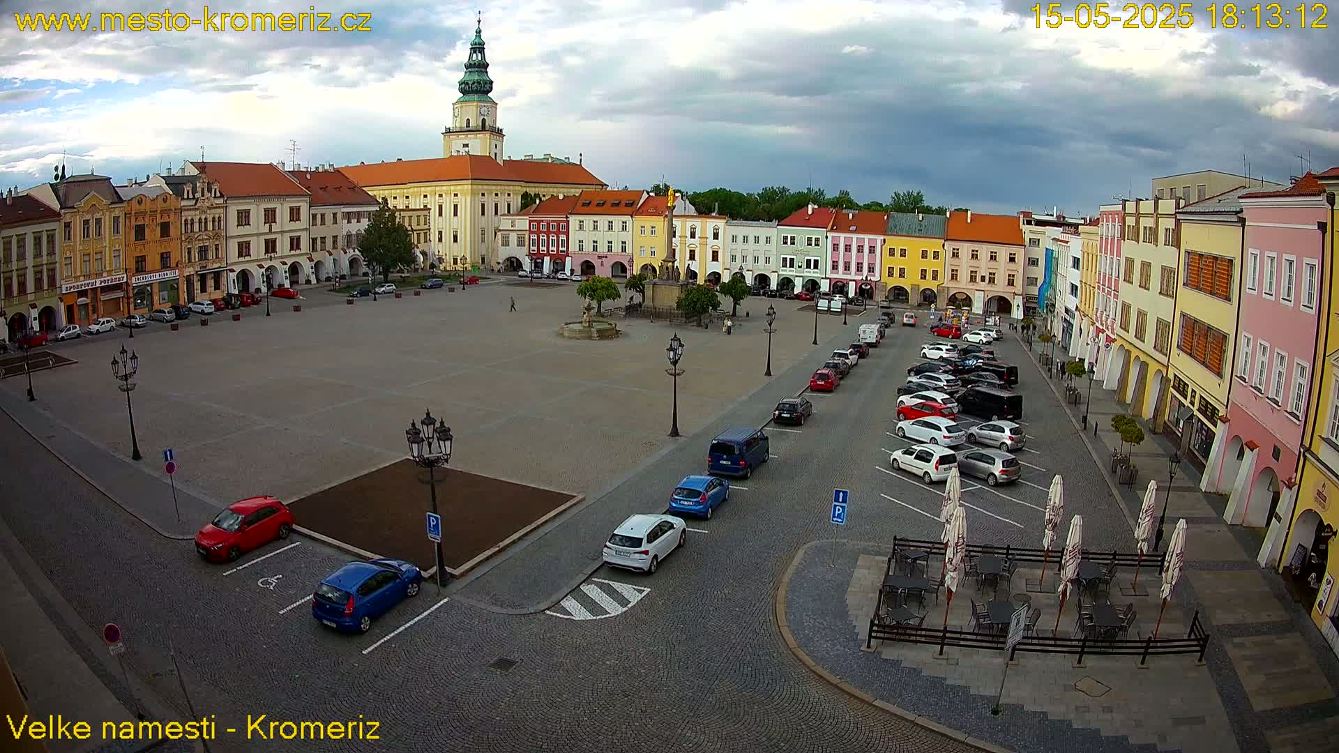 A large town square with colorful buildings surrounding it, some cars parked along the sides, and an outdoor cafe with umbrellas under a partly cloudy sky.