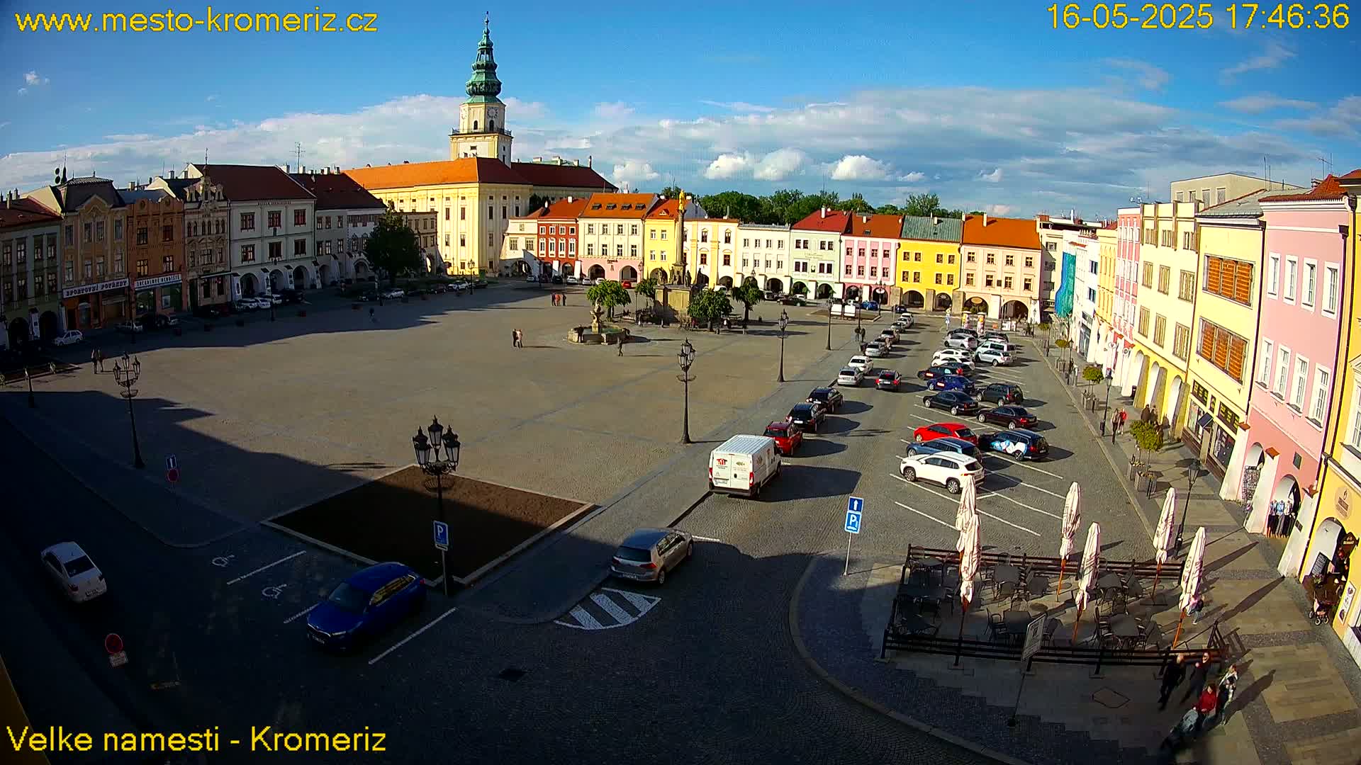 A large town square with colorful buildings, a fountain in the center, and parked cars under a mostly sunny sky.