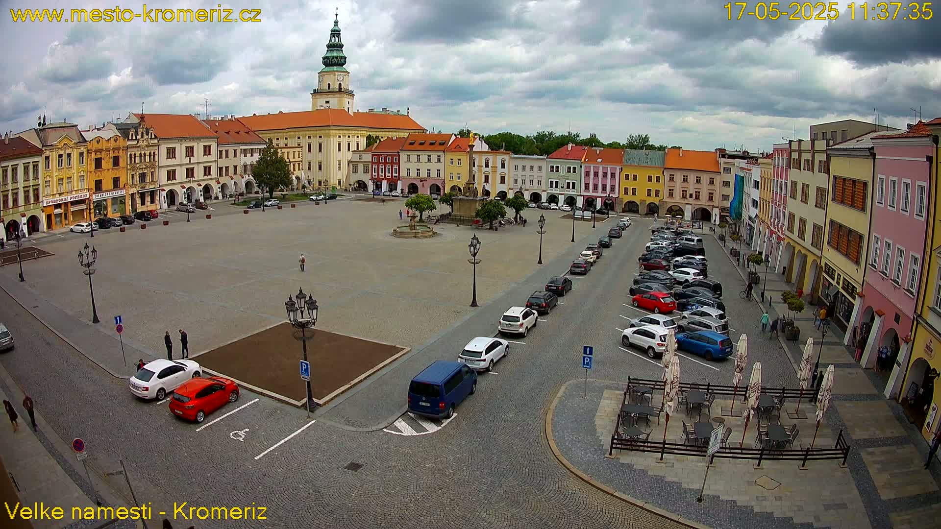 A large town square with colorful buildings surrounding it, under a partly cloudy sky, is filled with parked cars and a few pedestrians.