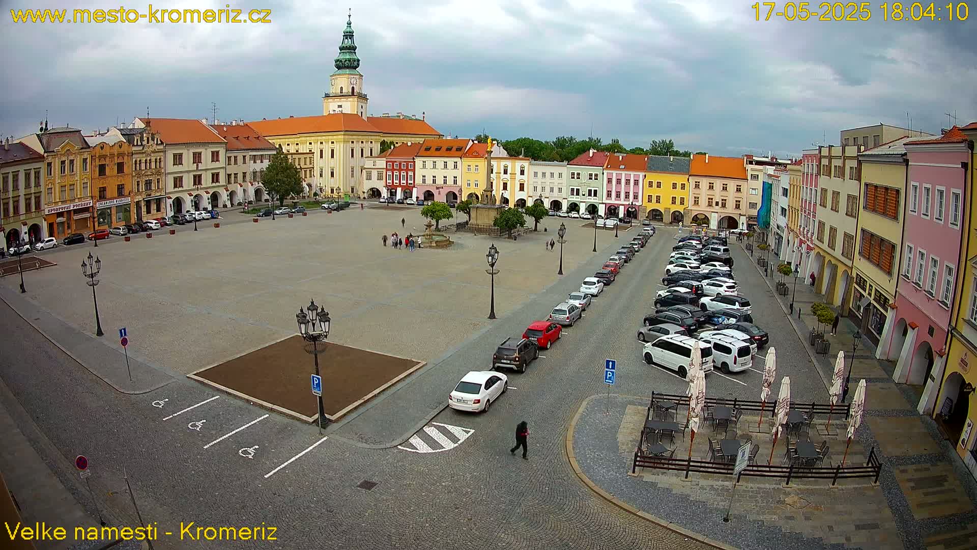 A large town square with colorful buildings surrounding it, a fountain in the center, cars parked along the sides, and overcast skies.
