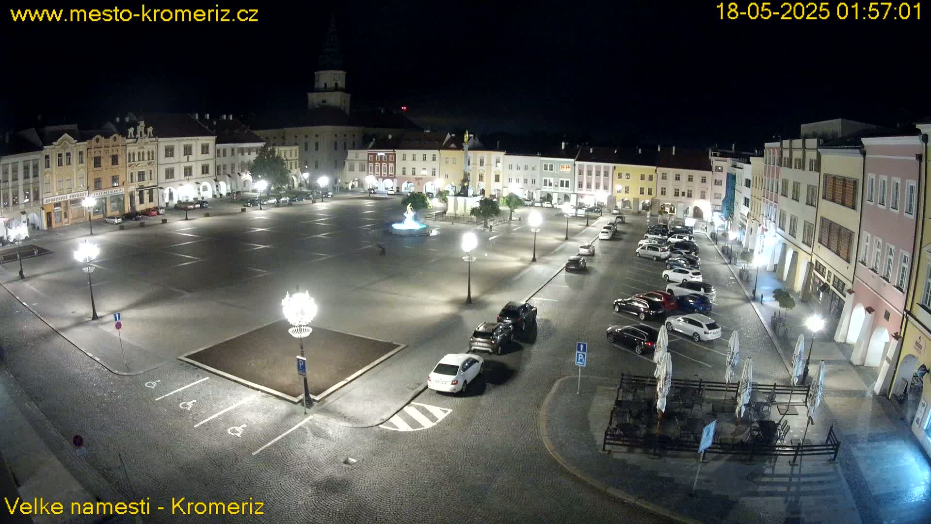 A nighttime view of a town square with several cars parked around the perimeter, a fountain in the center, and outdoor seating at a restaurant.