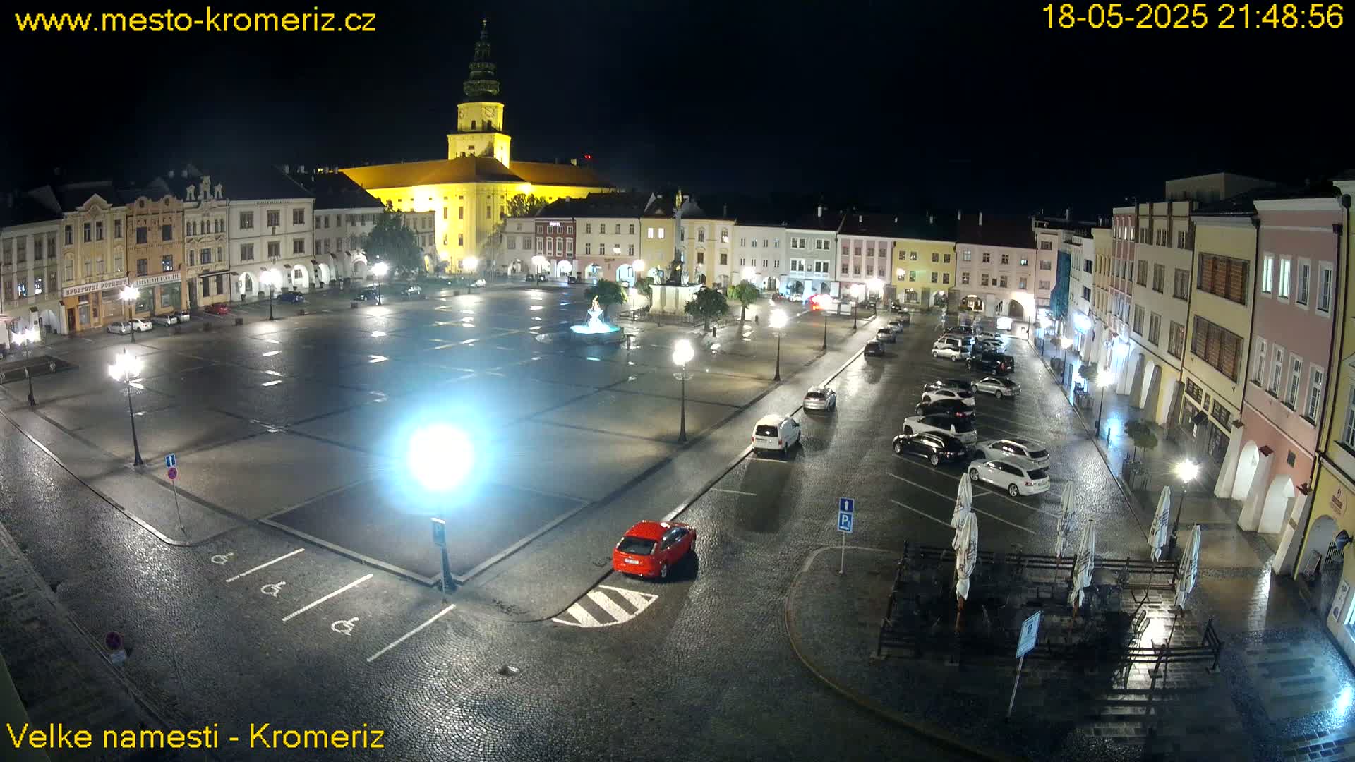 A rain-slicked town square at night, illuminated by streetlights, shows several cars parked and a few buildings surrounding the open space.