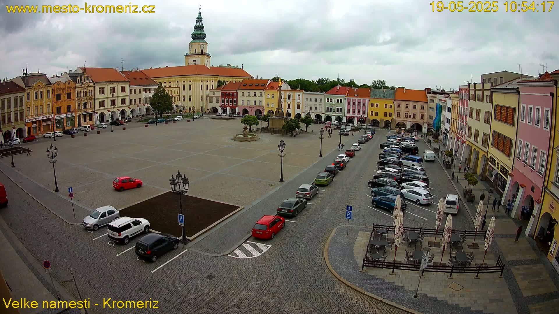 A large town square with colorful buildings surrounding it, partially filled with parked cars under an overcast sky.