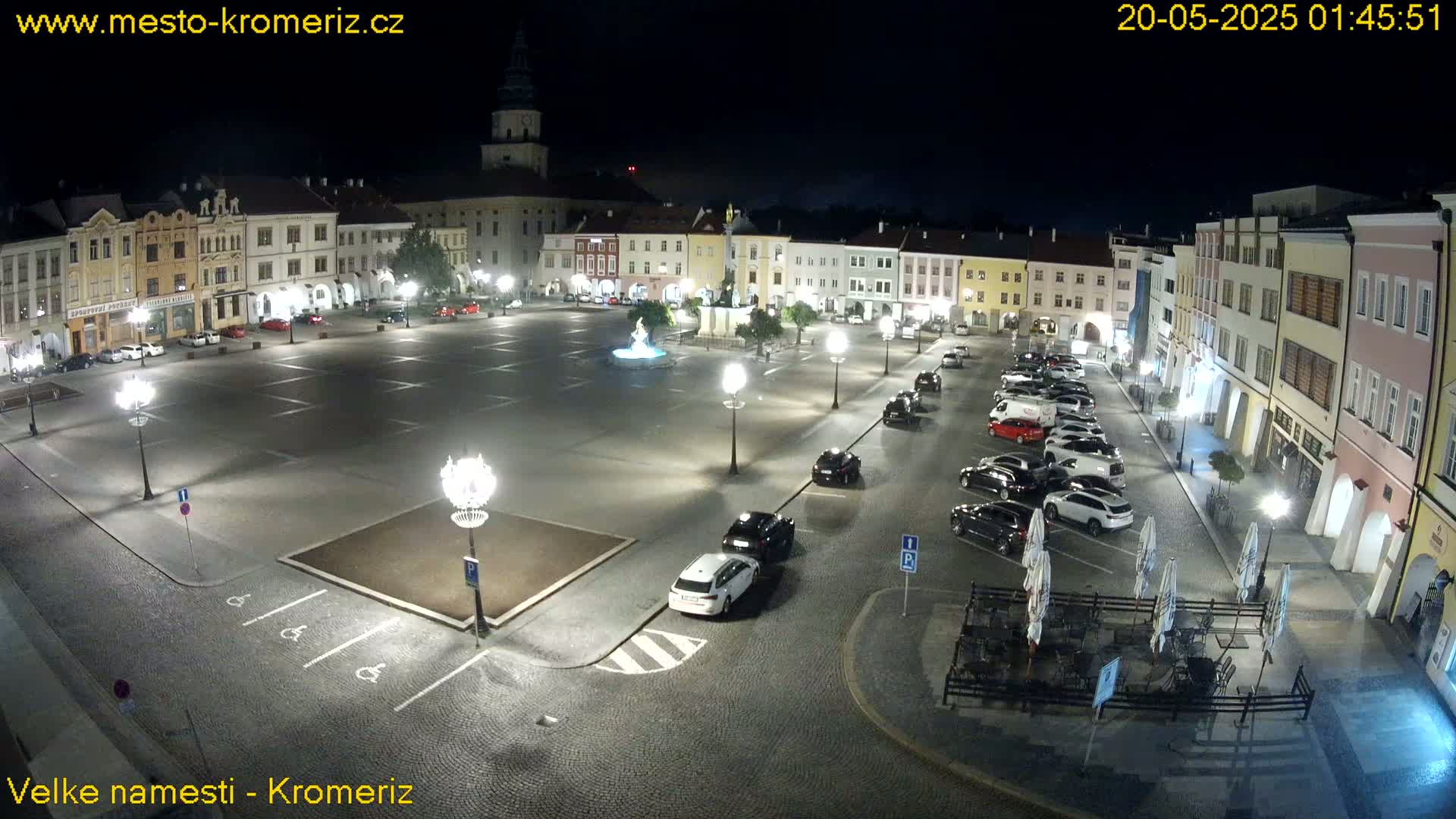 A nighttime view of a town square with several cars parked, buildings illuminated, and a fountain at its center under clear skies.