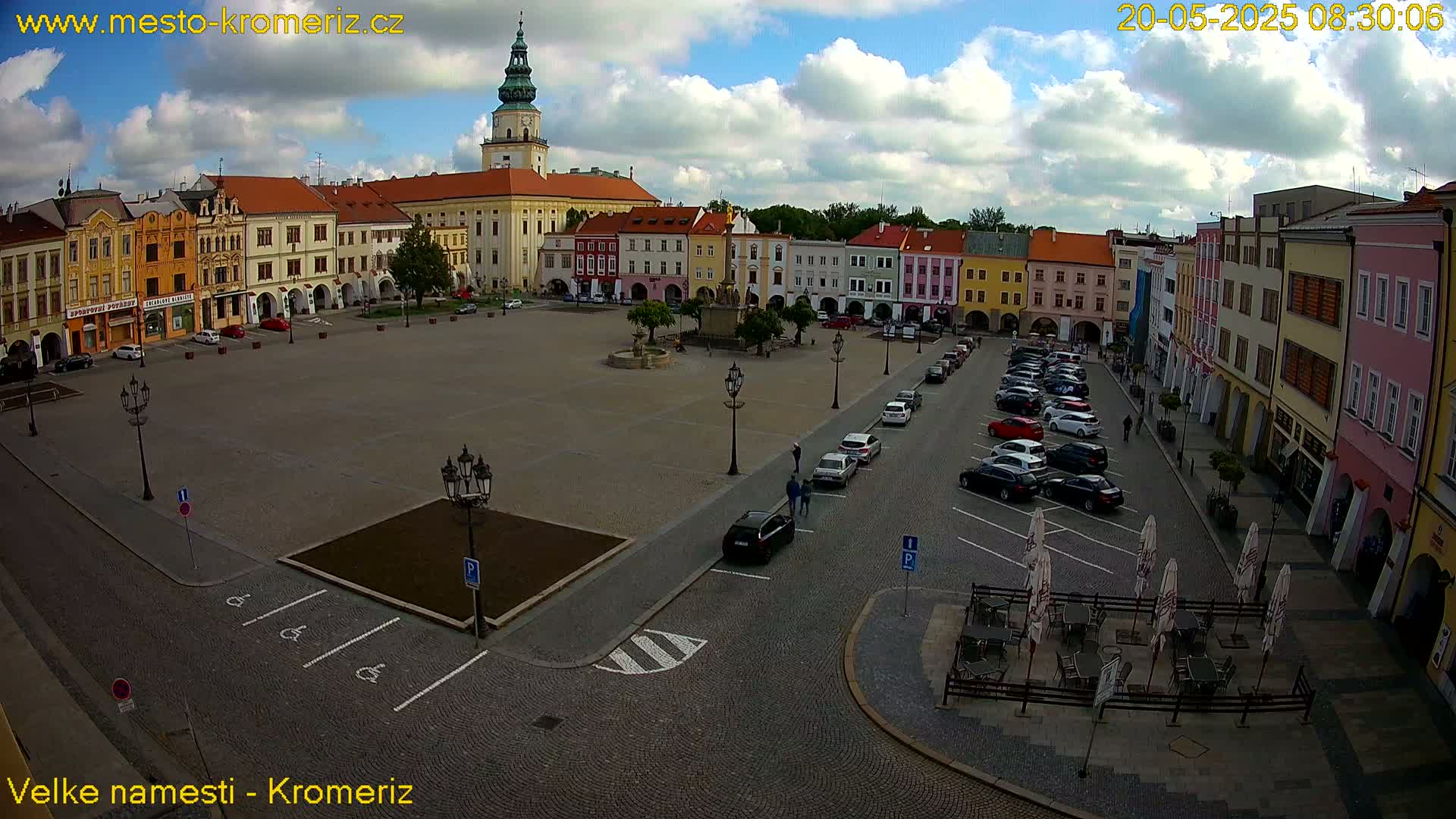 A large town square with colorful buildings surrounding it, under a partly cloudy sky, is shown with several cars parked around the perimeter and some outdoor seating.