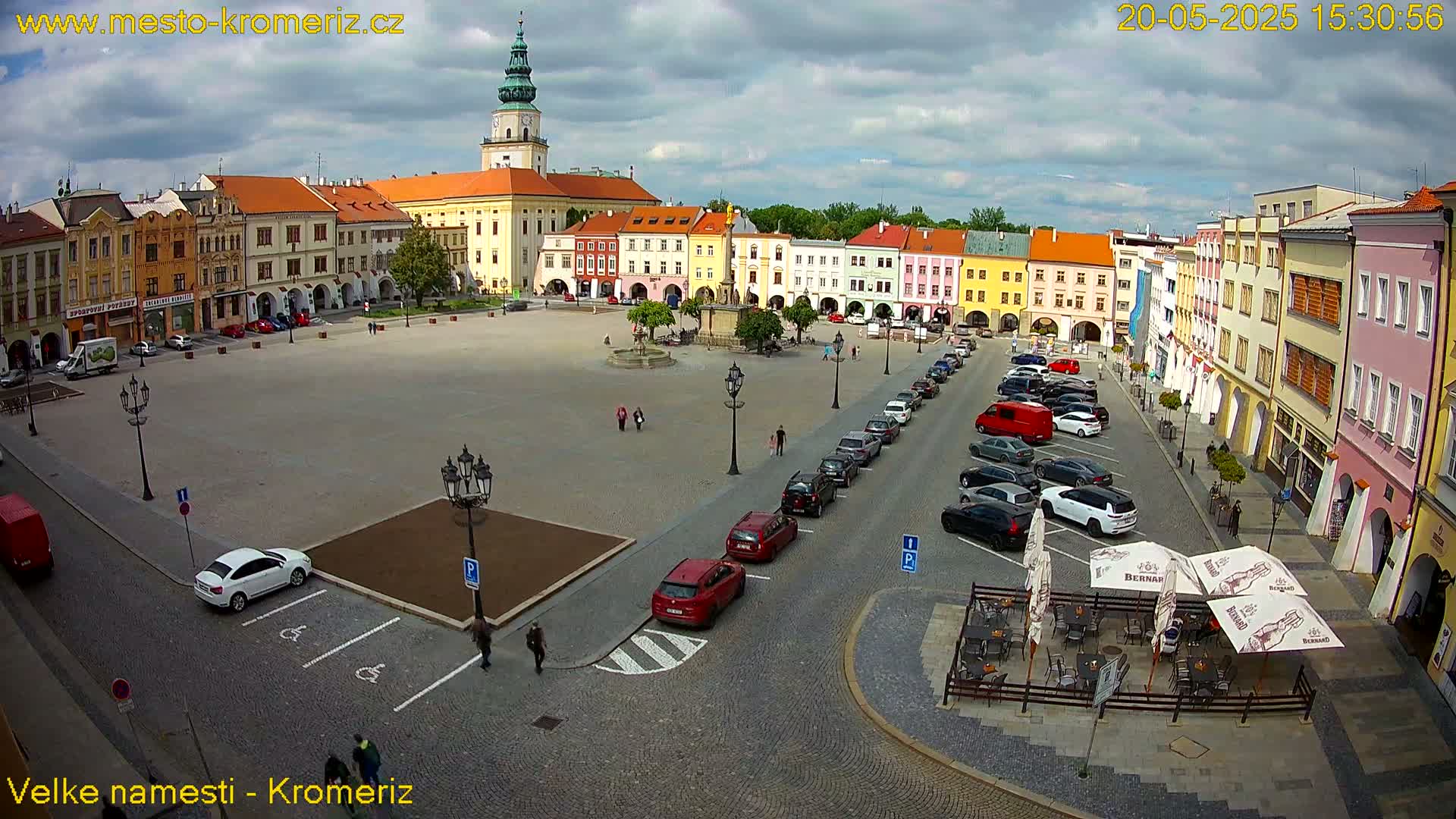 A large town square with colorful buildings surrounding it, some cars parked, and a few pedestrians, under a partly cloudy sky.