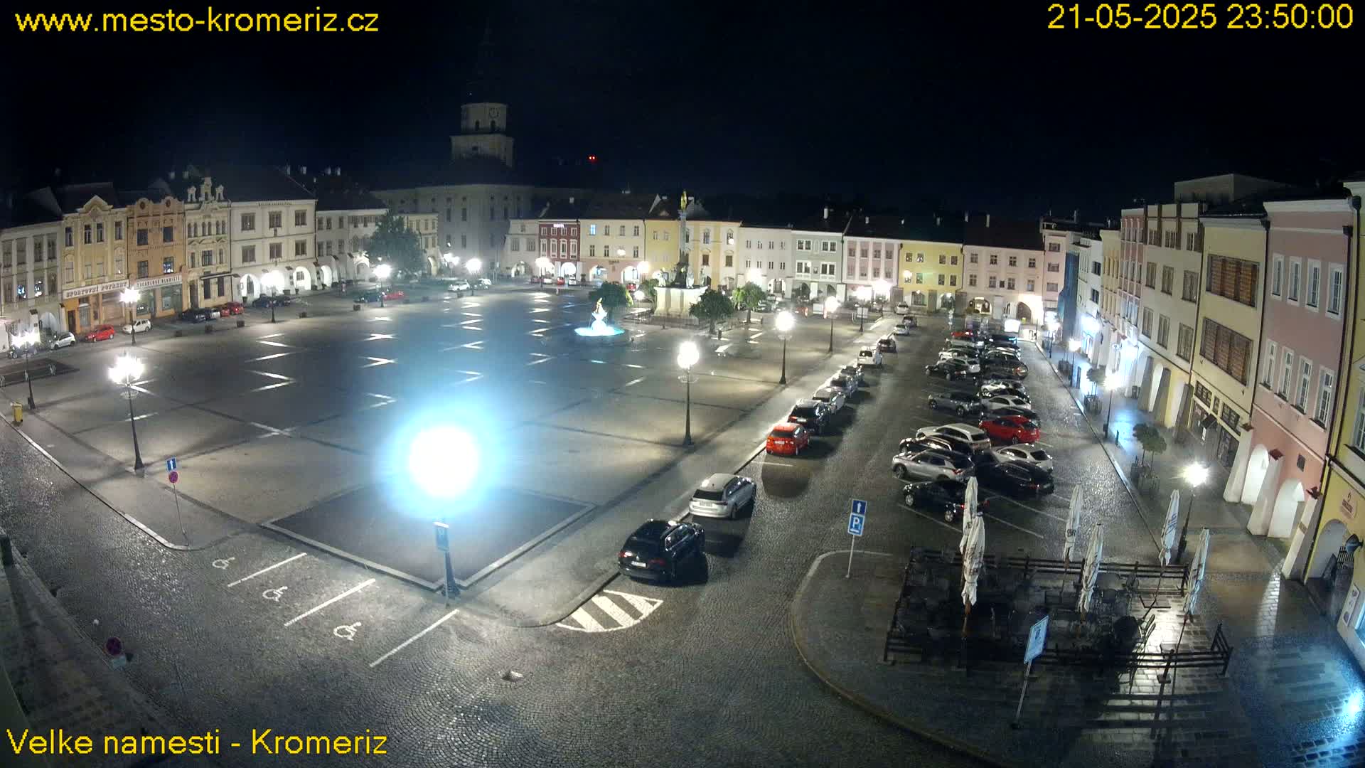 A nighttime view of a town square with cars parked along the sides and a few lights illuminating the area, under a slightly wet or rainy sky.