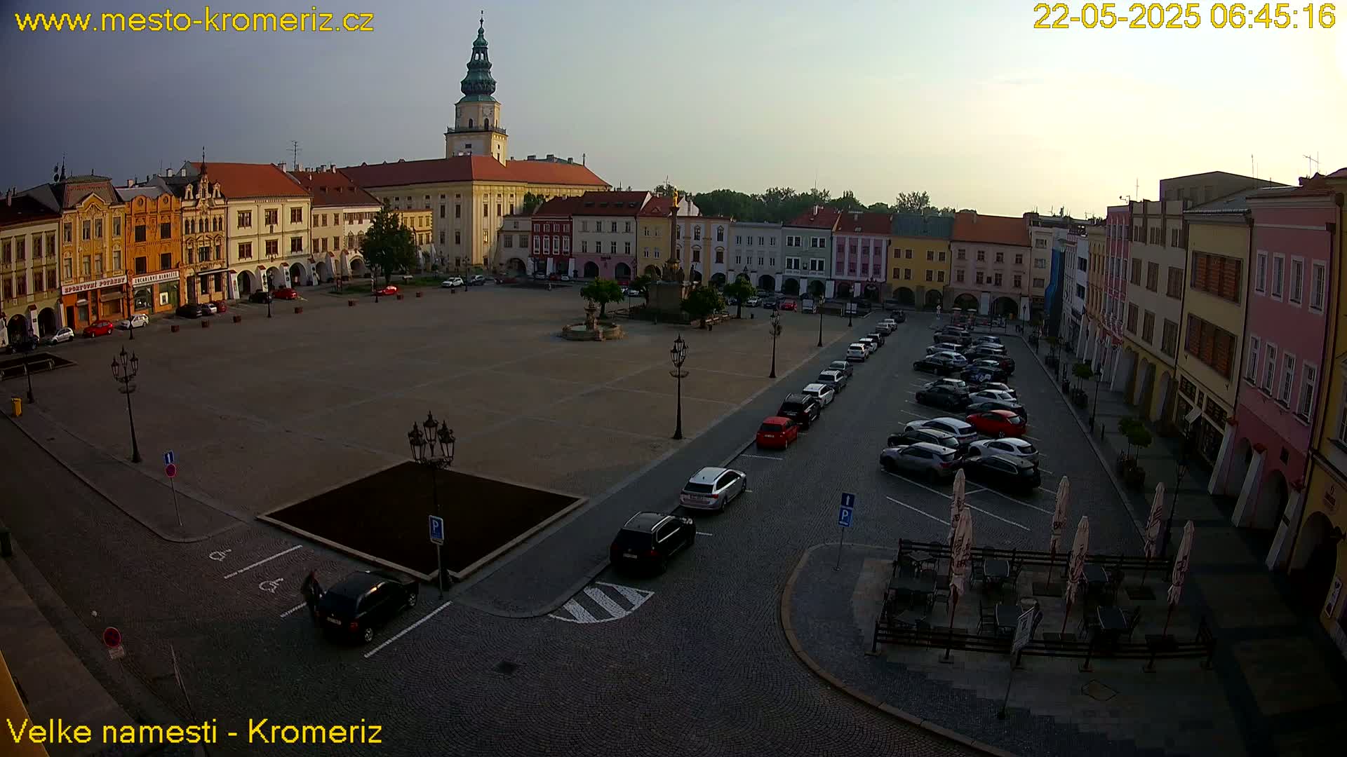 An expansive town square, surrounded by colorful buildings and a central fountain, is mostly empty under a clear, dusky sky with cars parked along the sides.