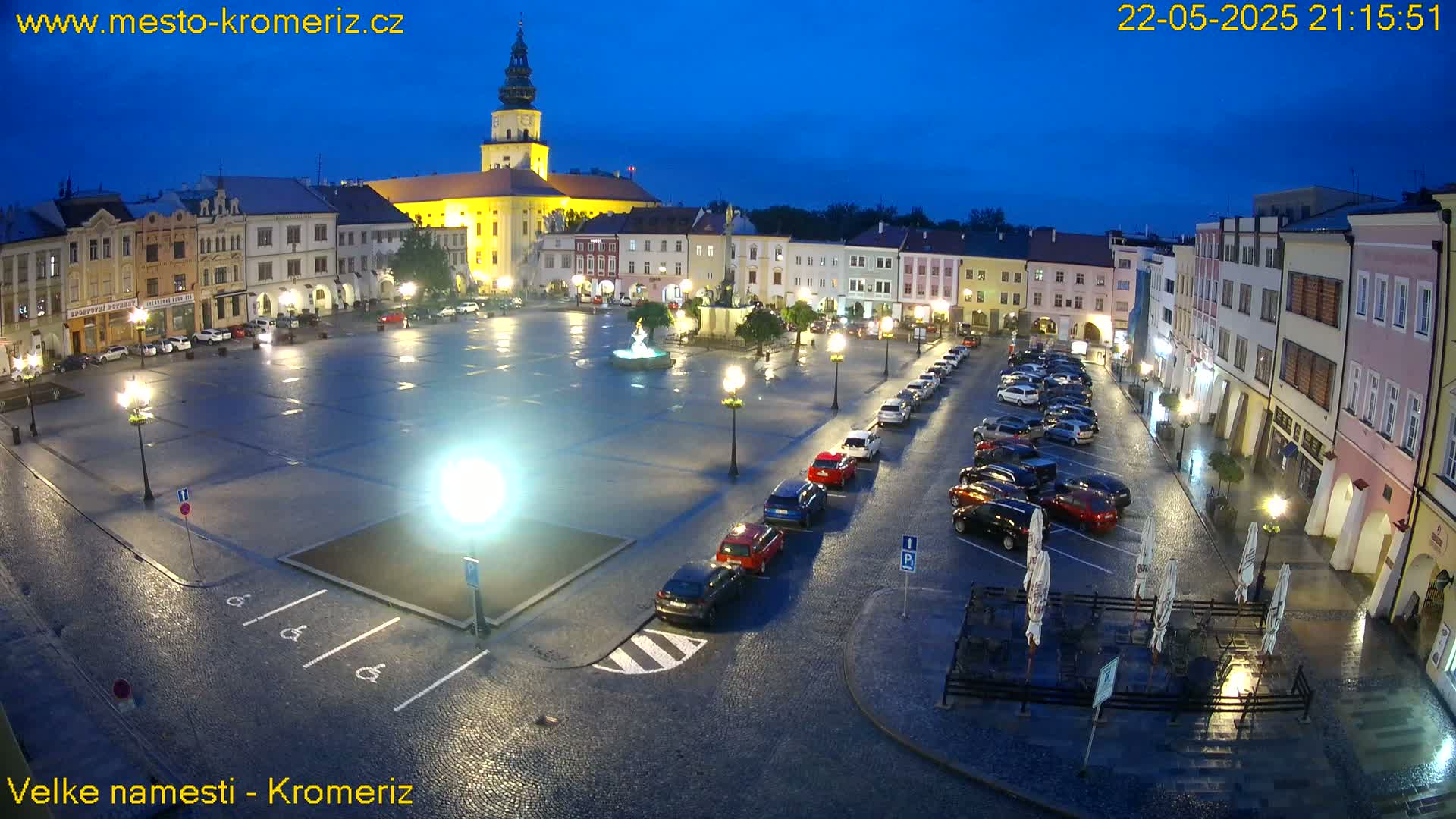 A town square at night, illuminated by streetlights, shows wet cobblestones, parked cars, and buildings surrounding a central fountain.