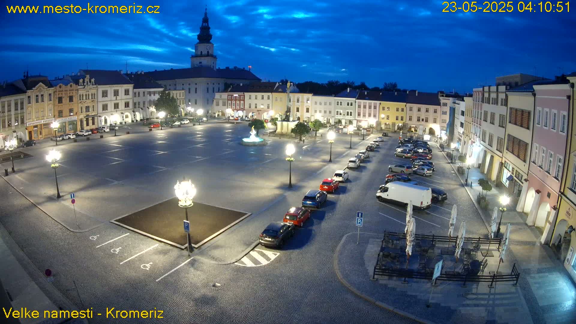 A large, mostly empty town square at twilight, with cars parked along the edges and a few outdoor tables and chairs set up near buildings, under a partly cloudy sky.