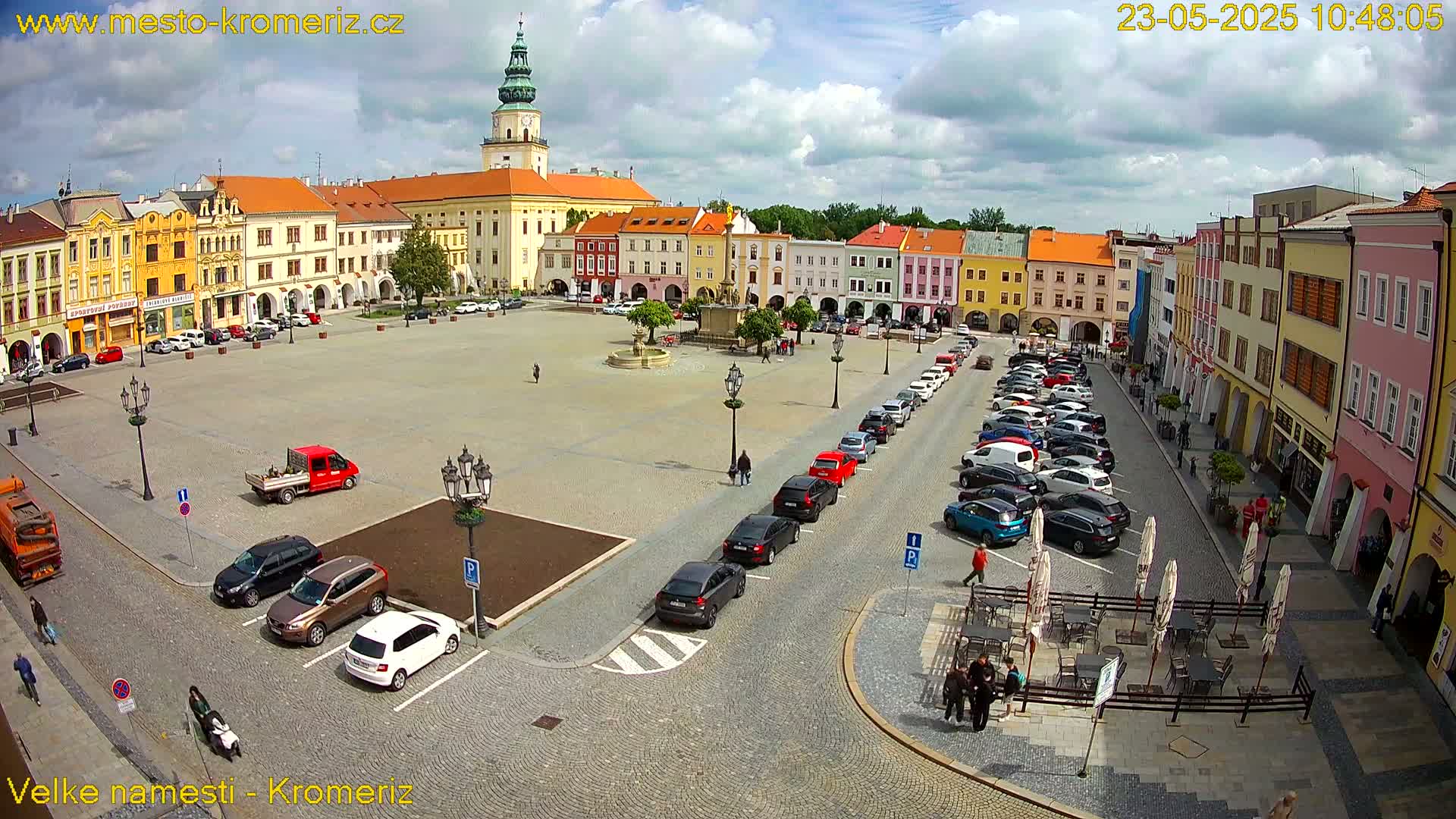 A large town square with colorful buildings surrounding it, under partly cloudy skies, has cars parked along the sides and a few people walking around.