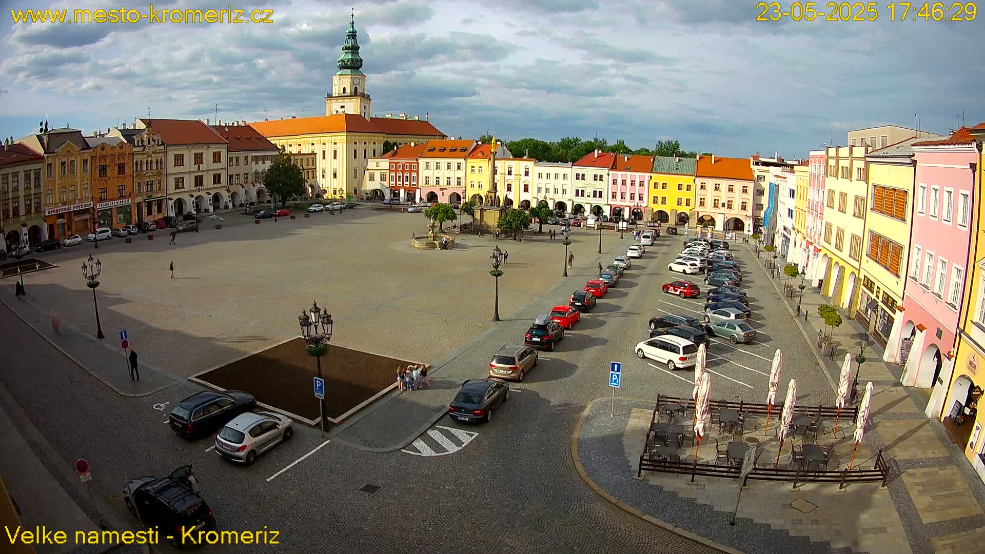 A large town square with colorful buildings surrounding it, cars parked along the sides, and a few people walking around under partly cloudy skies.