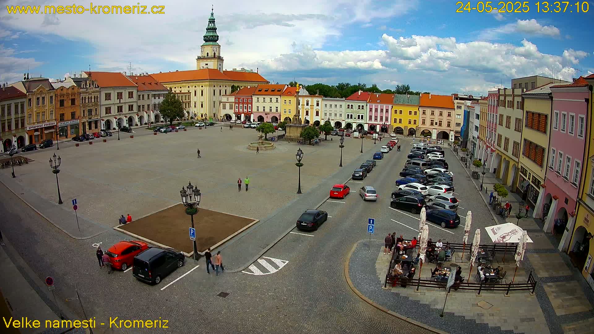 A town square with colorful buildings surrounding a paved area, containing parked cars, a few people, and outdoor seating, under a partly cloudy sky.