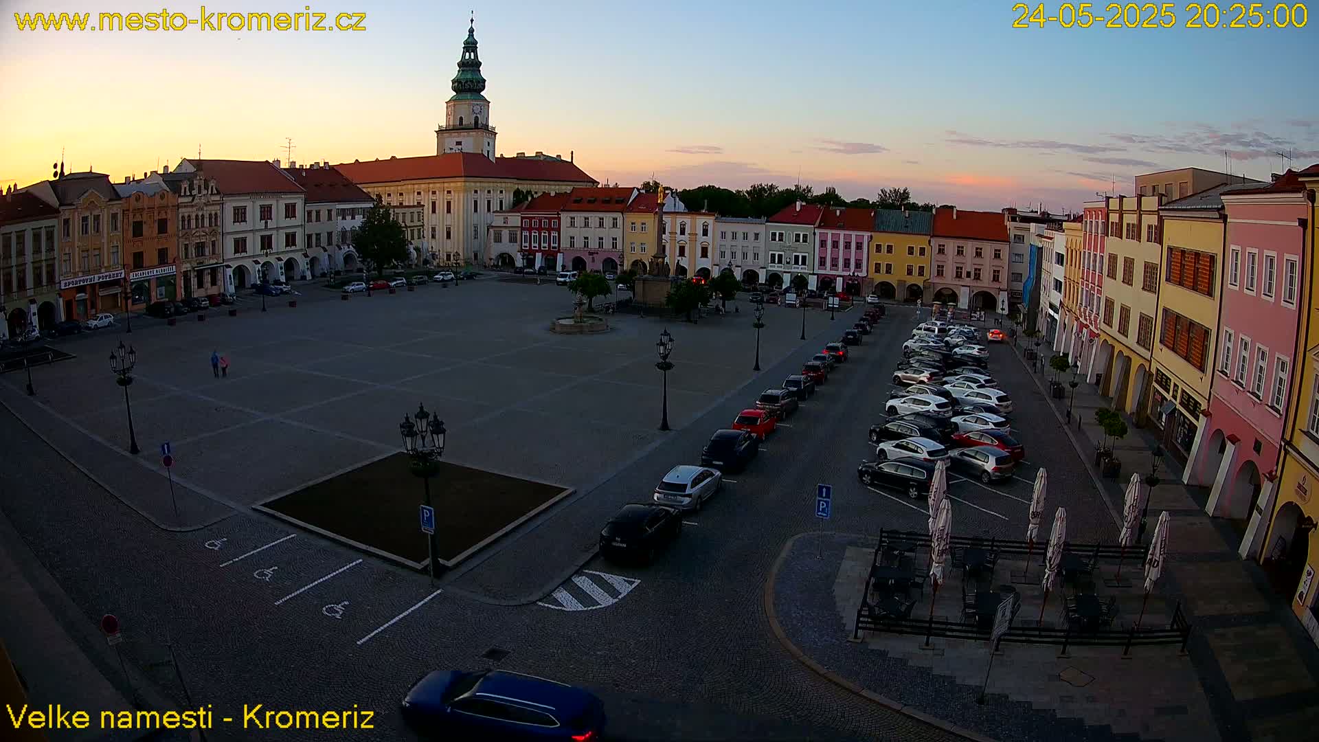 A large town square with colorful buildings surrounding it, partially filled with parked cars under a twilight sky.