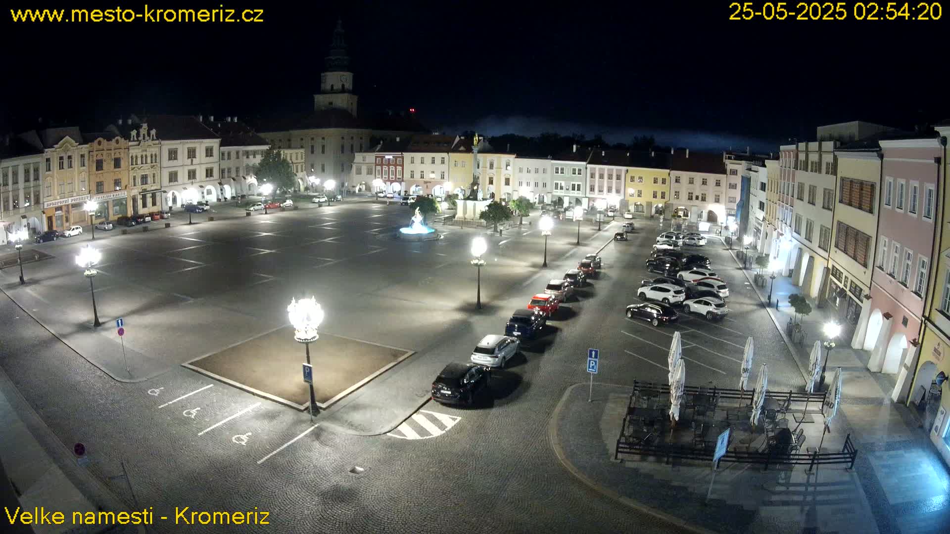 A town square at night, illuminated by streetlights, with several cars parked along the edges and a fountain in the center, under a slightly hazy sky.