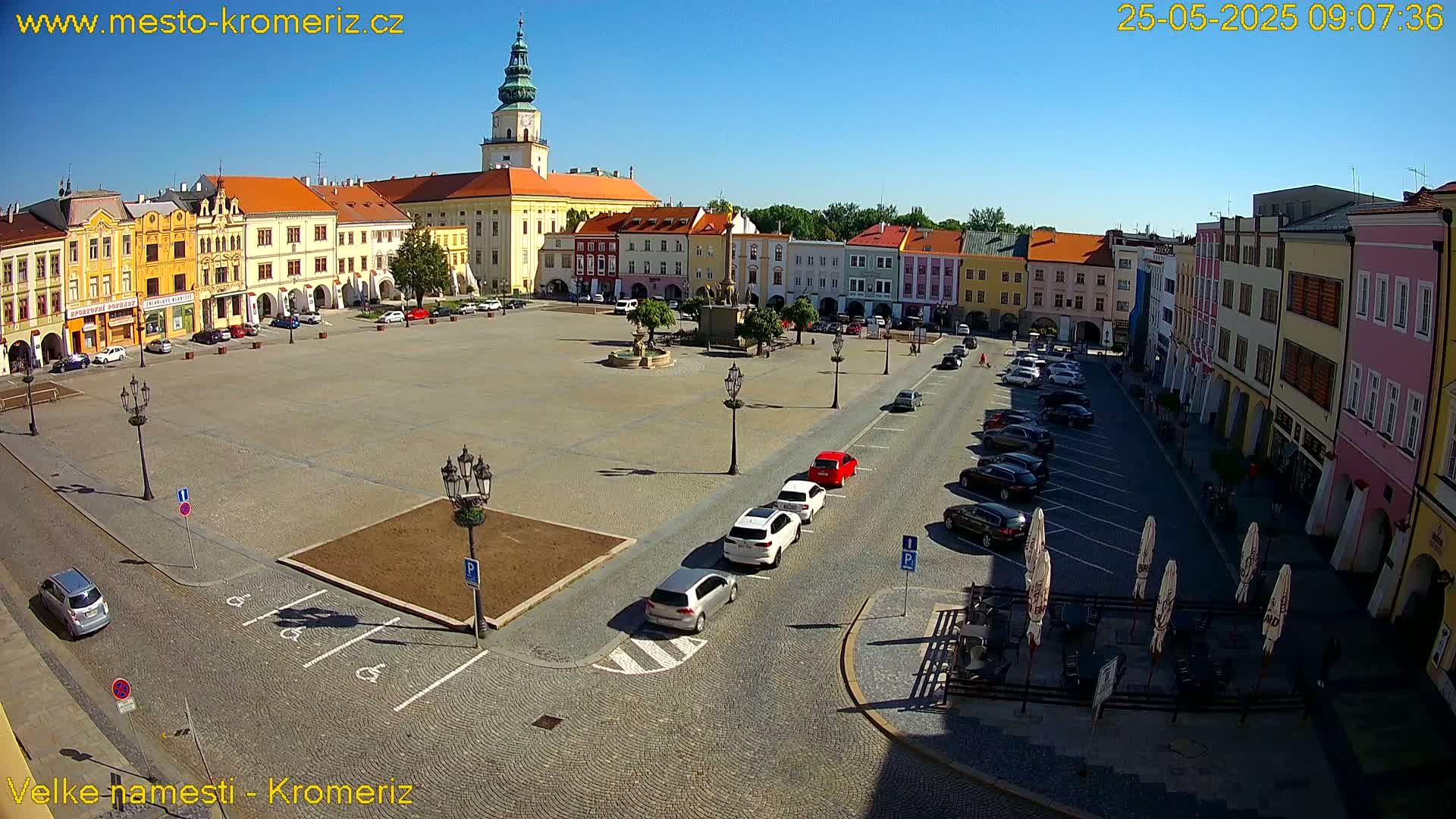 A large town square with colorful buildings surrounding it, cars parked along the sides, and a few cars driving on the square itself under a clear, sunny sky.