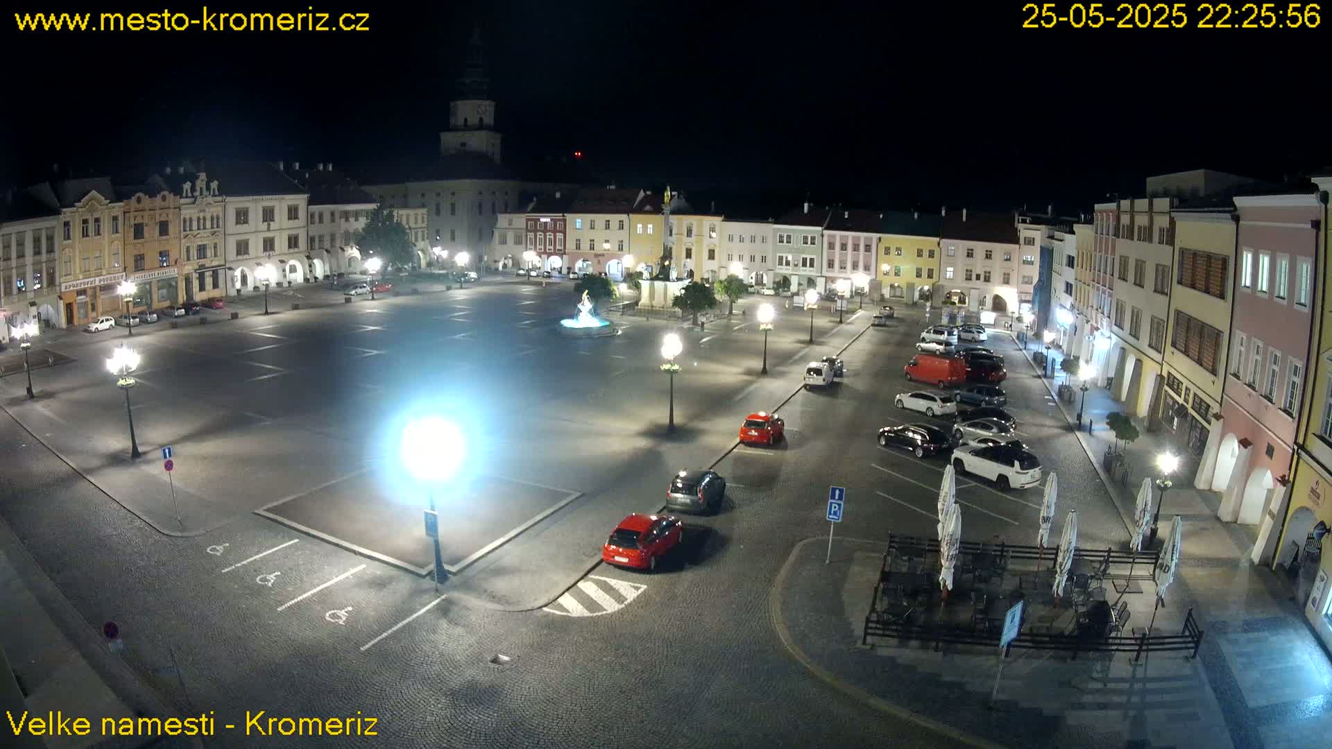 A nighttime, illuminated town square with parked cars and a few outdoor tables, under a clear sky.