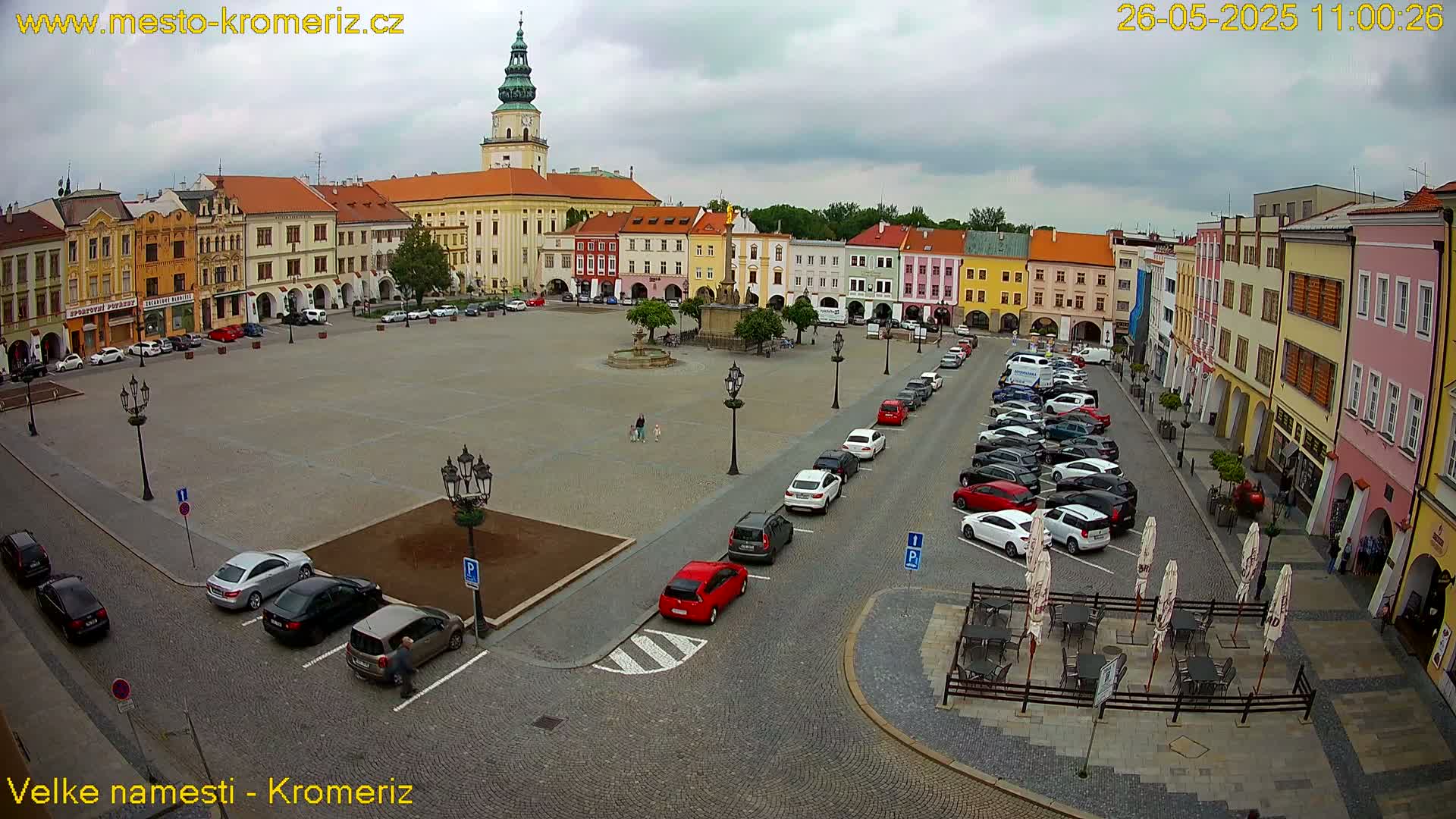 A large town square with colorful buildings surrounding it, many cars parked along the sides, and a few people walking around under an overcast sky.