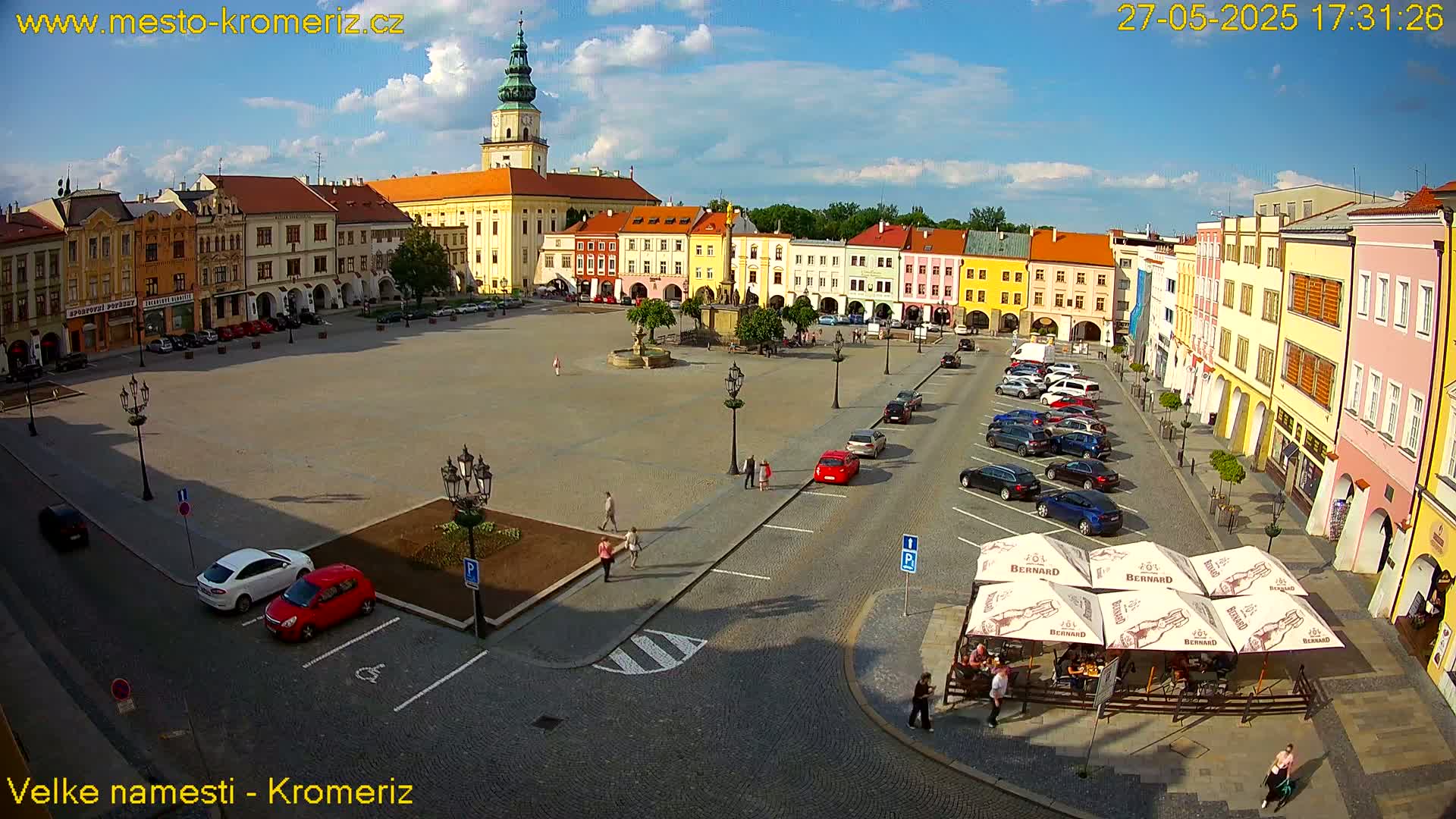 A large town square with colorful buildings surrounding it, under a mostly sunny sky with a few clouds, is partially filled with parked cars and a few pedestrians.
