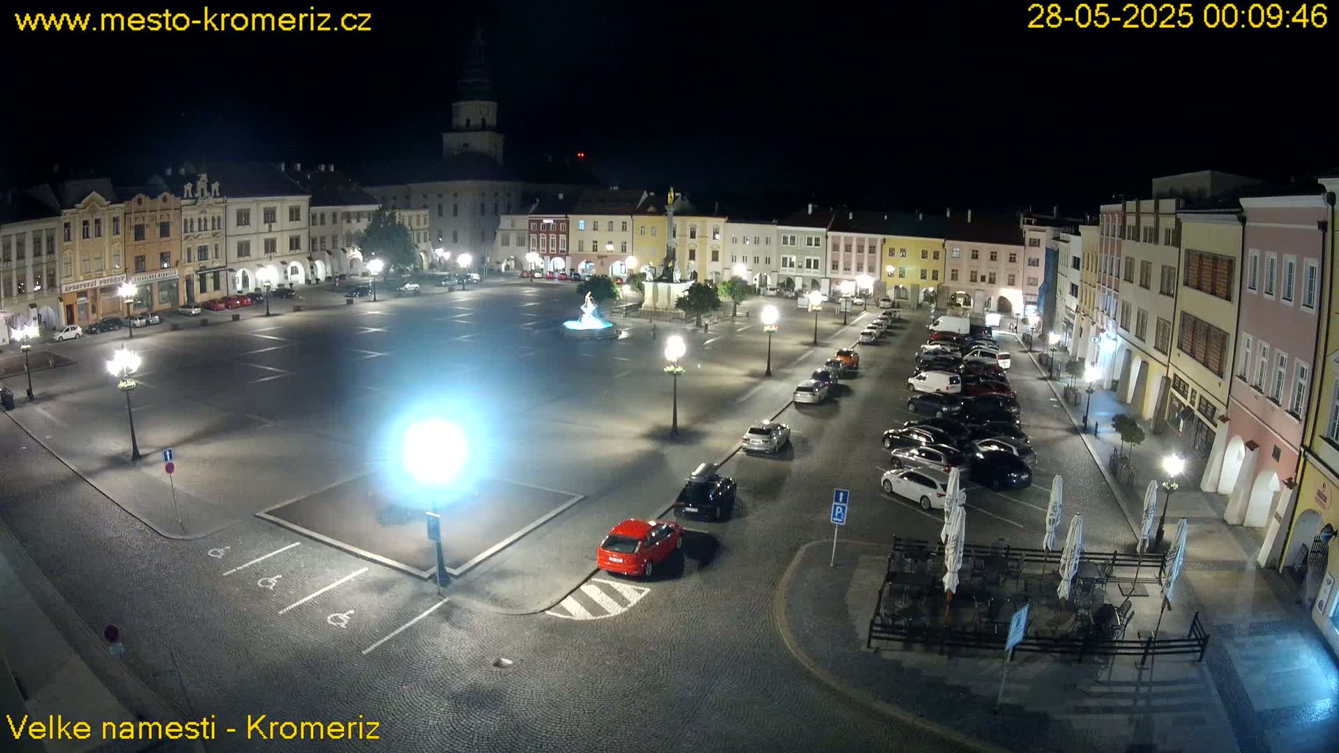 A nighttime, illuminated town square with cars parked along the sides and a fountain in the center.