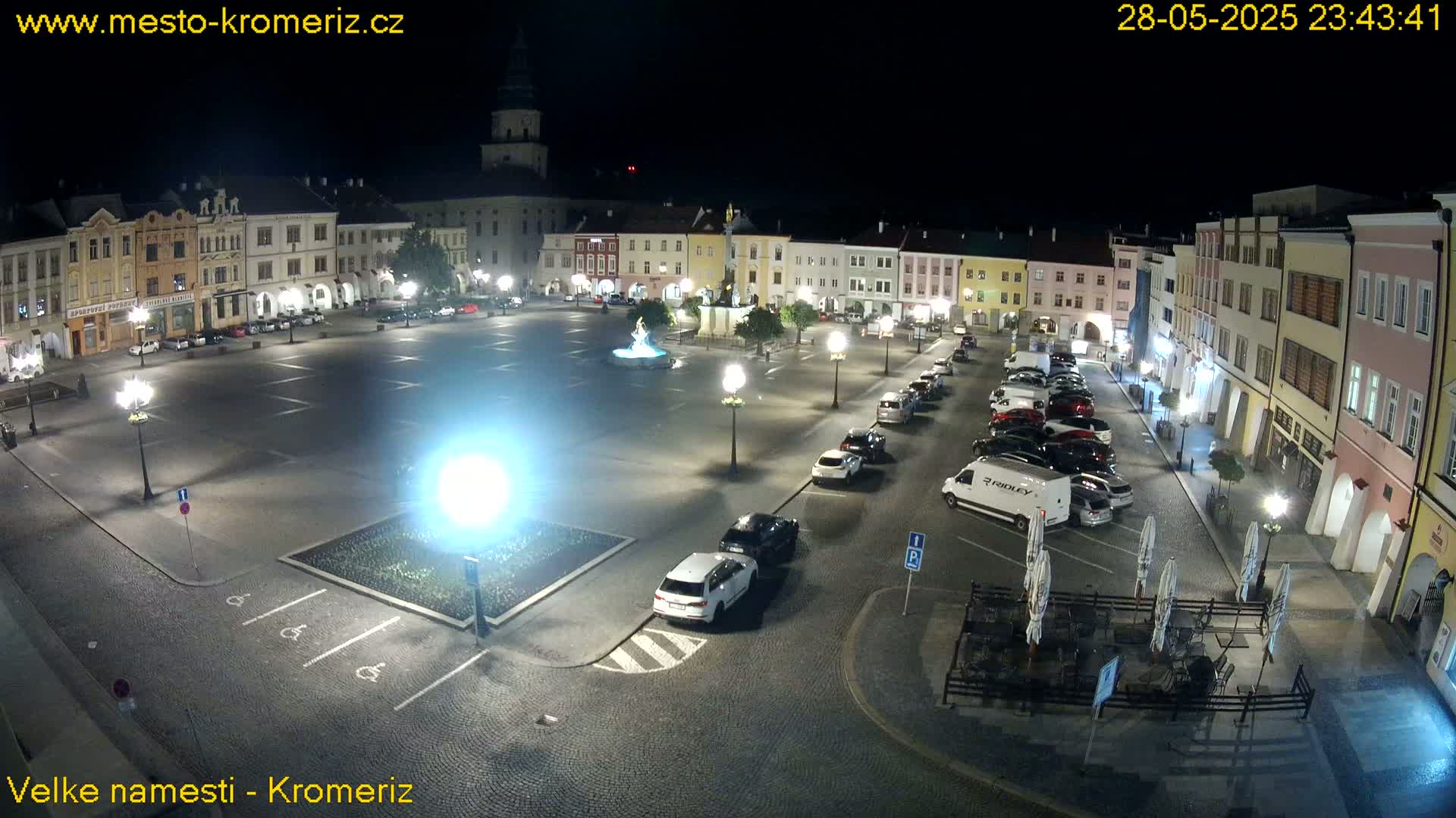 A nighttime, brightly lit town square with parked cars and a fountain, surrounded by buildings.